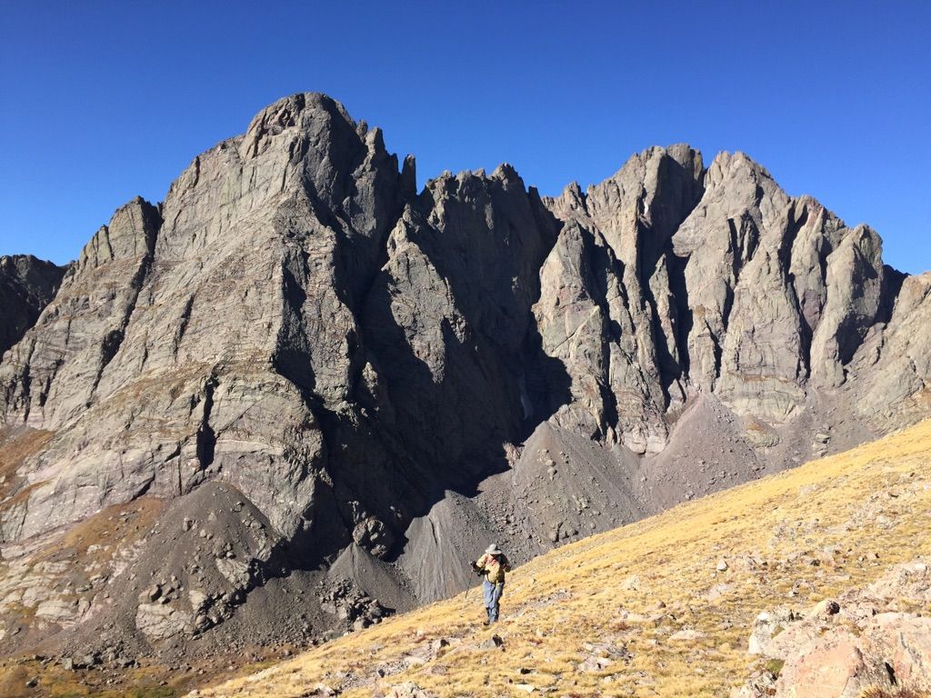 A person walks up a golden grassy slope towards massive rocky mountains. - Humboldt Peak Trail