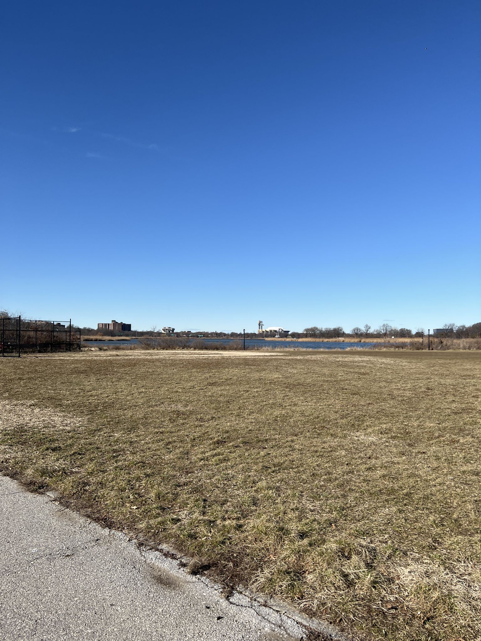 Wide, dry grassy field stretching towards distant water under a clear blue sky. - Brooklyn-Queens Greenway Loop