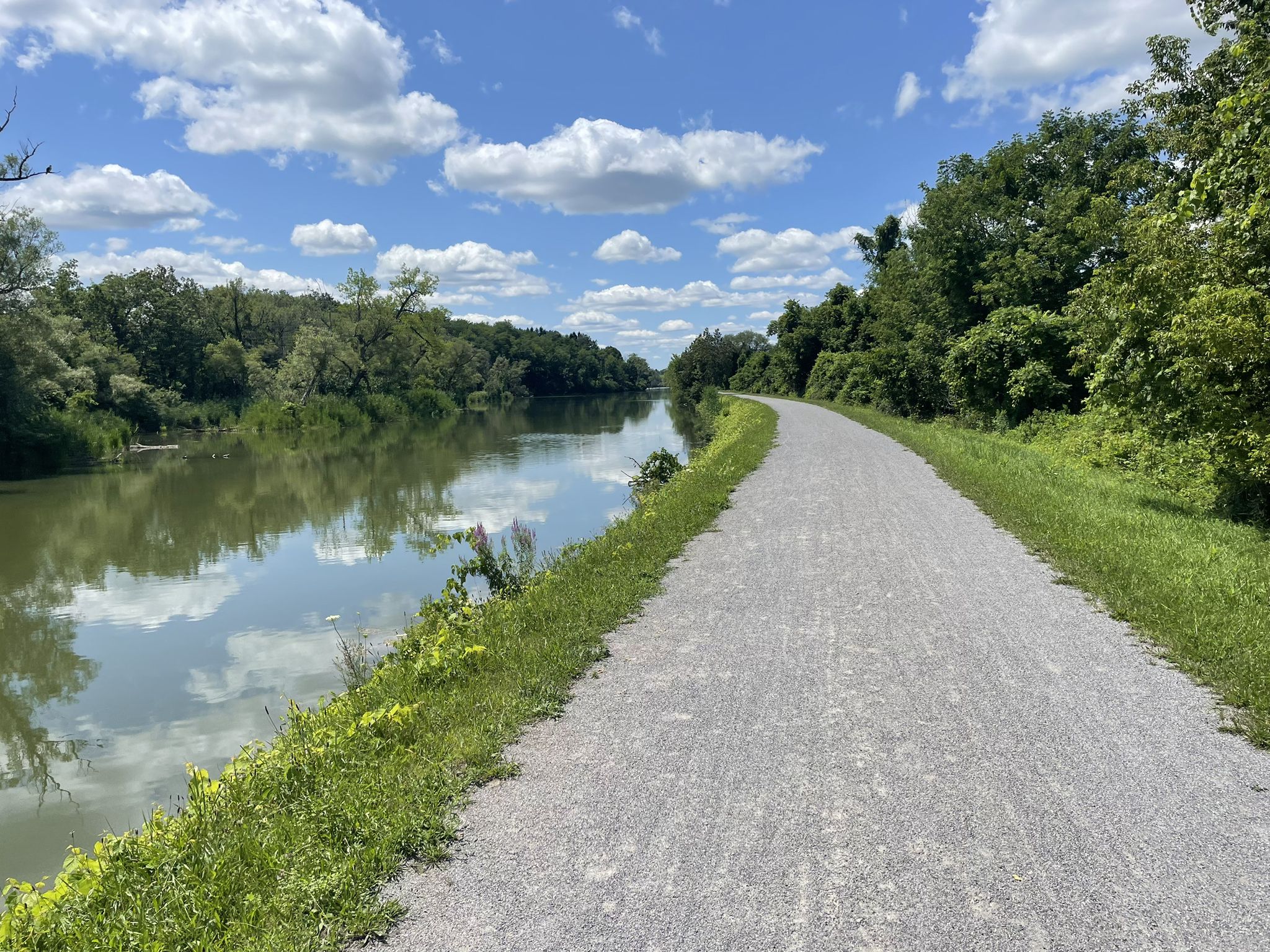 Gravel path alongside a calm waterway, surrounded by green trees under a sunny sky. - Erie Canalway Trail – Green Lake loop from Green Lakes State Park