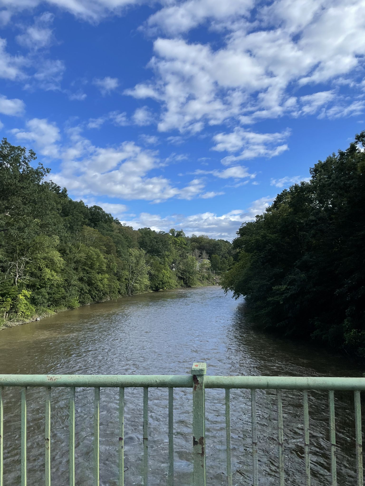 A river flows under a bridge, surrounded by green trees, under a blue sky. - Claverack Creek Waterfall – Albany-Hudson Electric Trail loop from Hudson