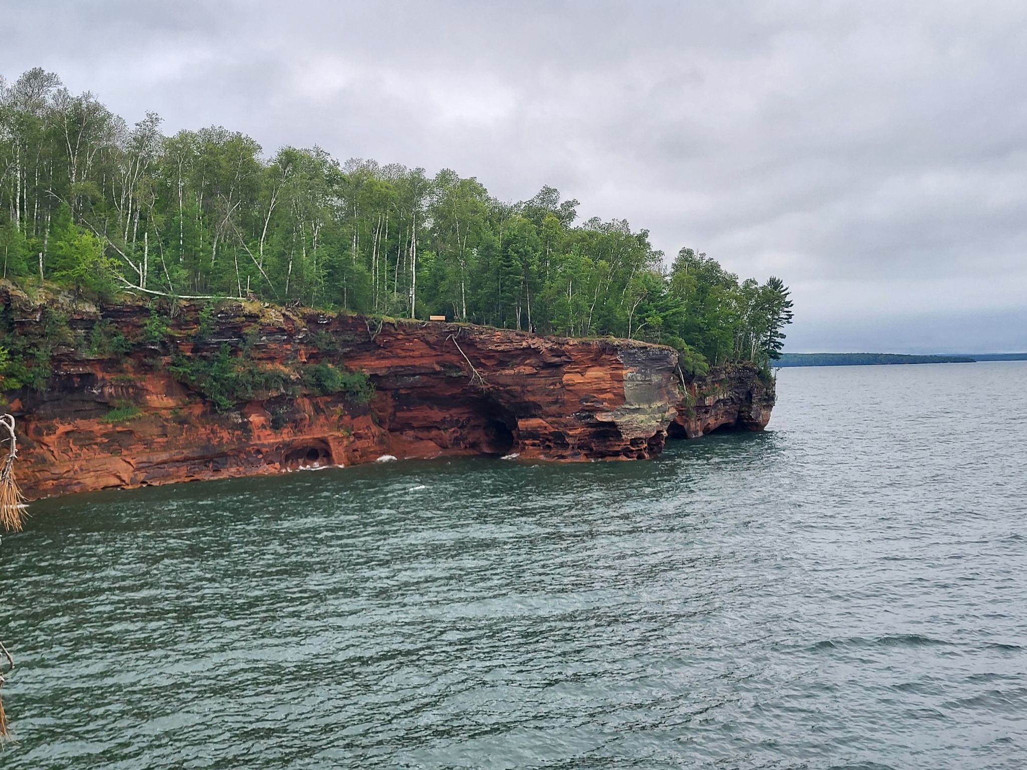 Red rock cliffs with green trees overlooking dark water under a cloudy sky. - Lakeshore Trail
