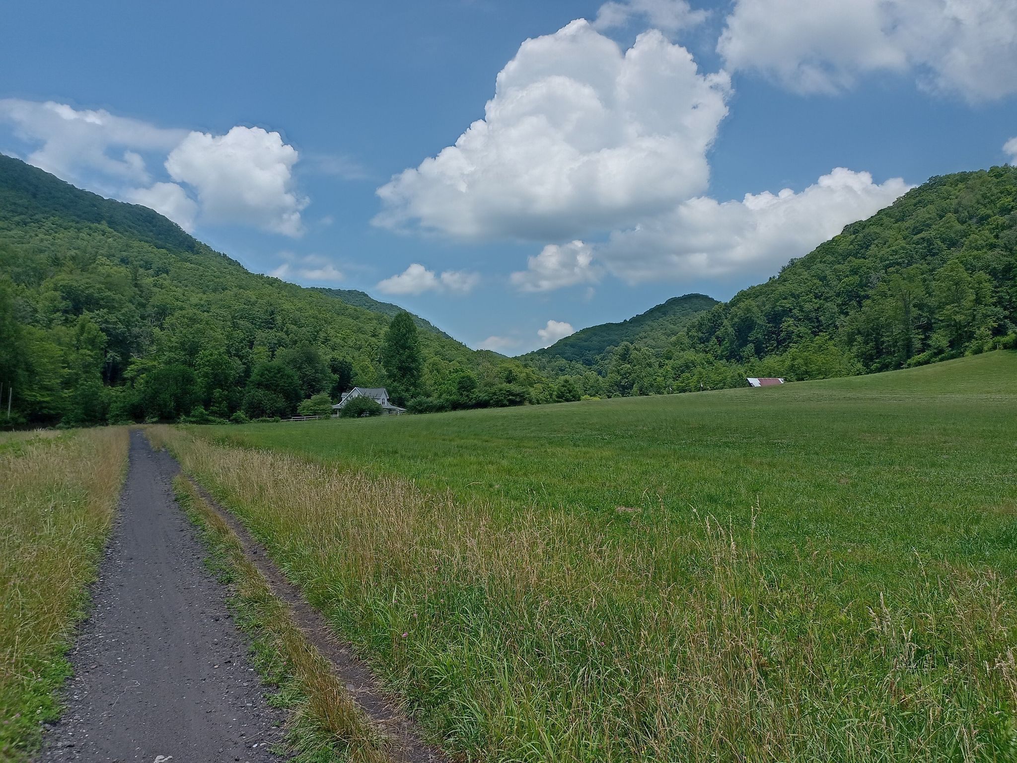 Dirt path through a sunny green valley with forested mountains and clouds. - Virginia Creeper Trail