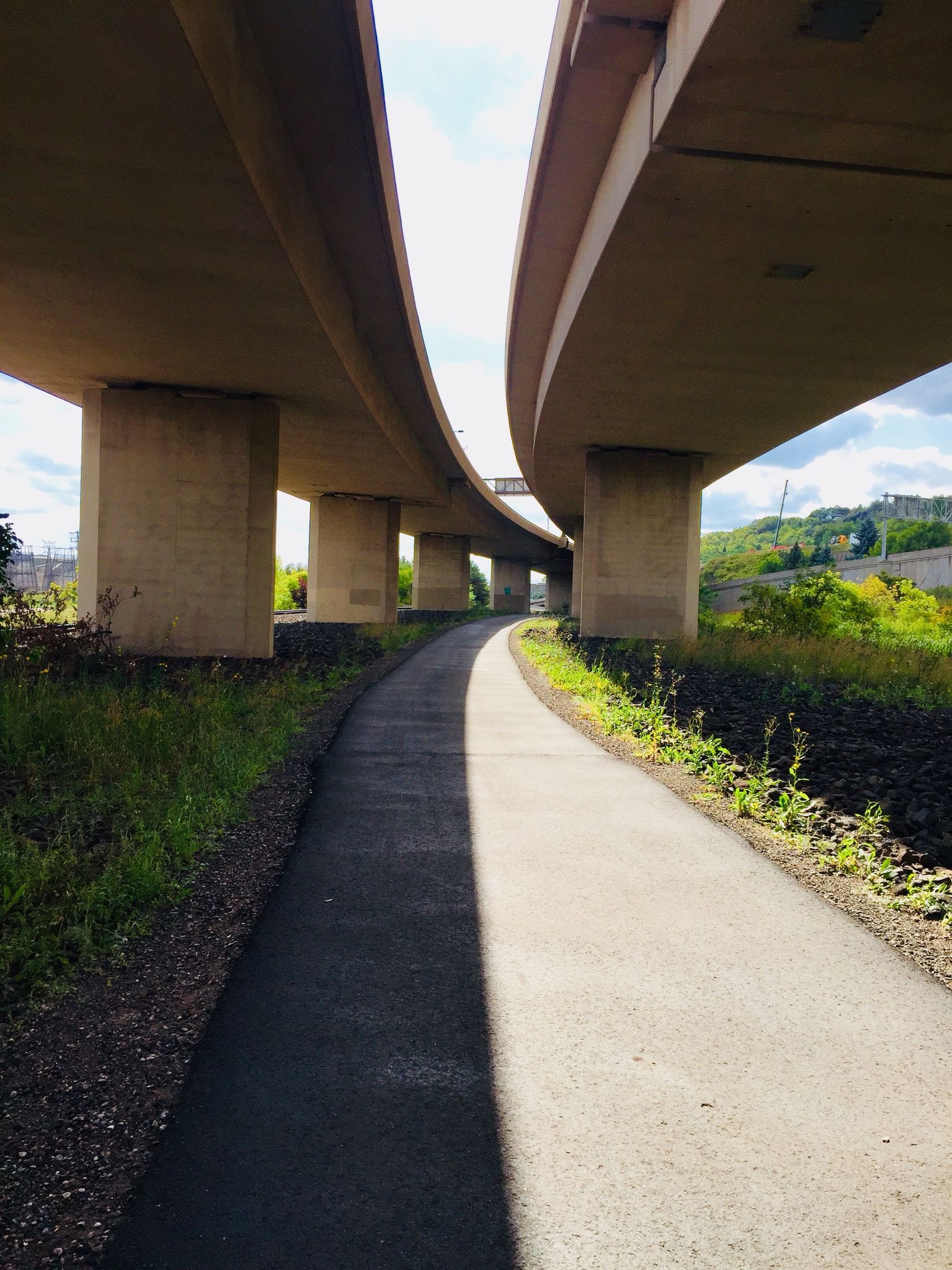 Paved path under concrete overpasses, bright sunlight creating strong shadows. - Lift Bridge to Enger Tower Trail