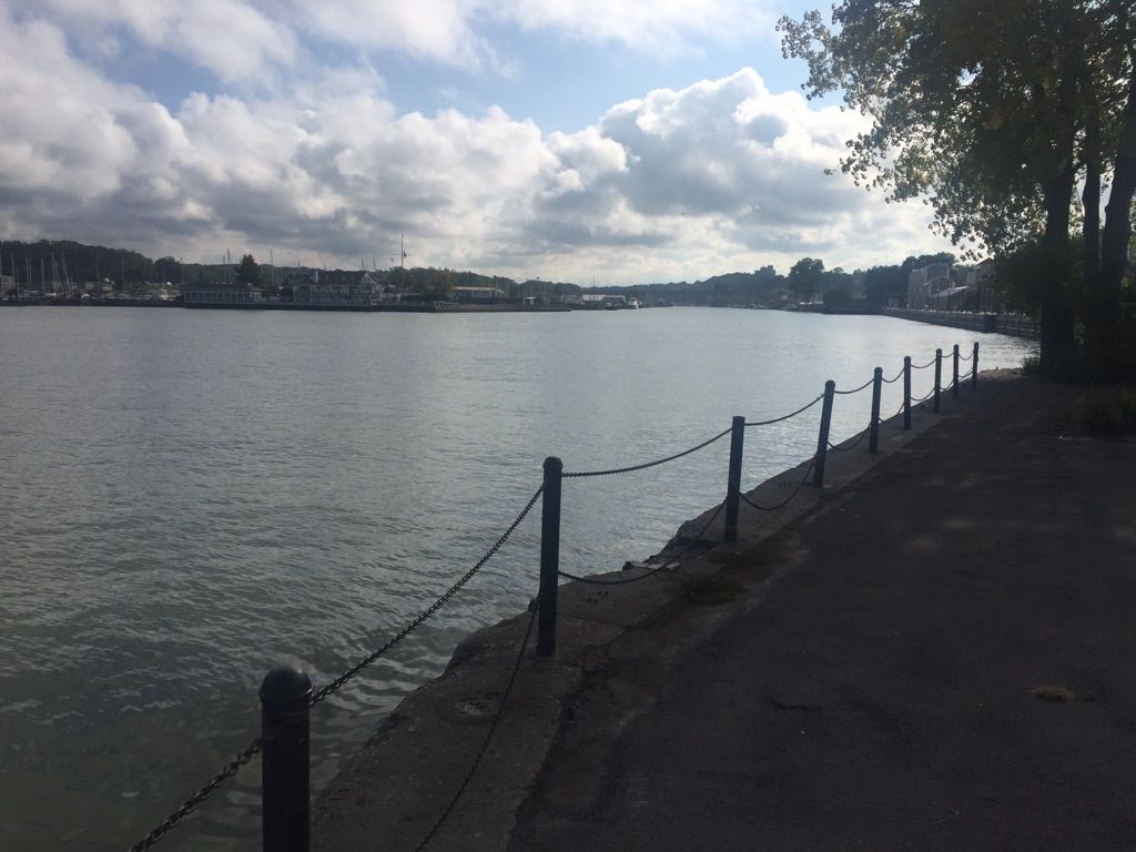 Cloudy day over wide water, with a paved path and distant town. - Charlotte–Genesee Lighthouse loop from Charlotte