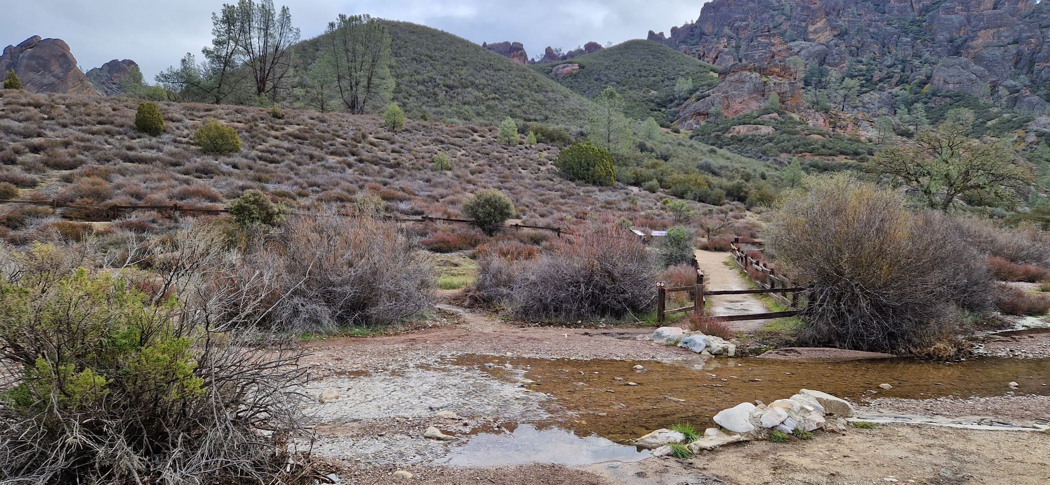 Stream flowing through dry, scrubby hills with rocky peaks under overcast sky. - North Wilderness Trail to Twin Knolls