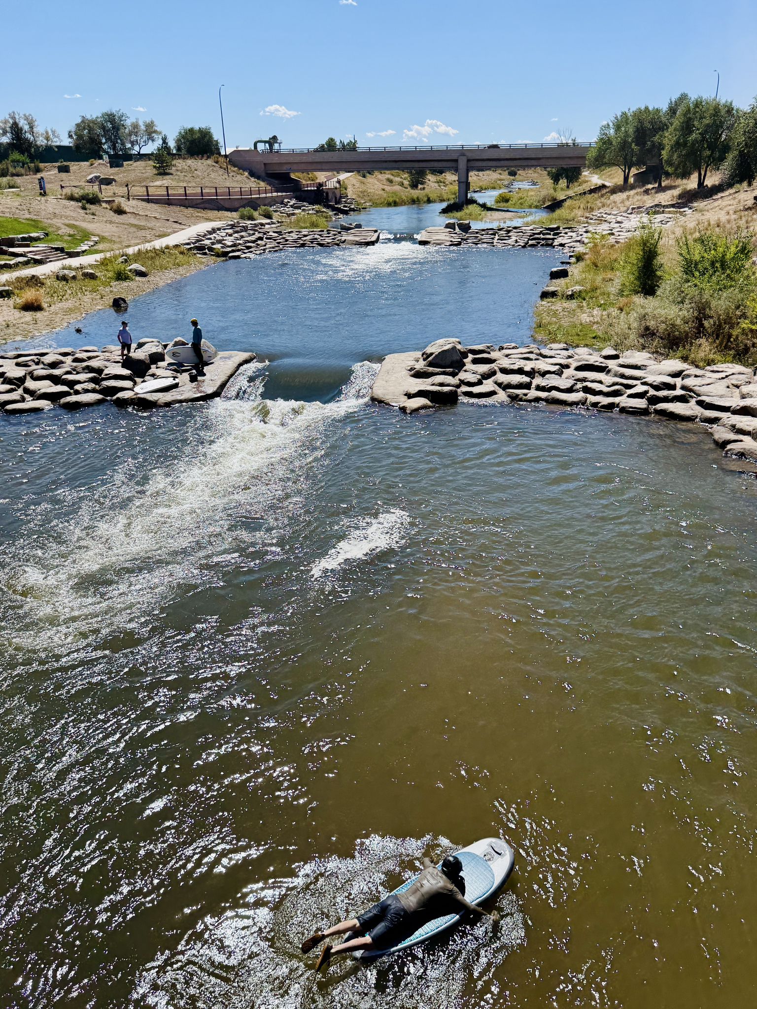 A person paddleboarding in a river with a small waterfall and bridge. - Bear Creek Lake – Bridge over South Platte River loop from Morrison