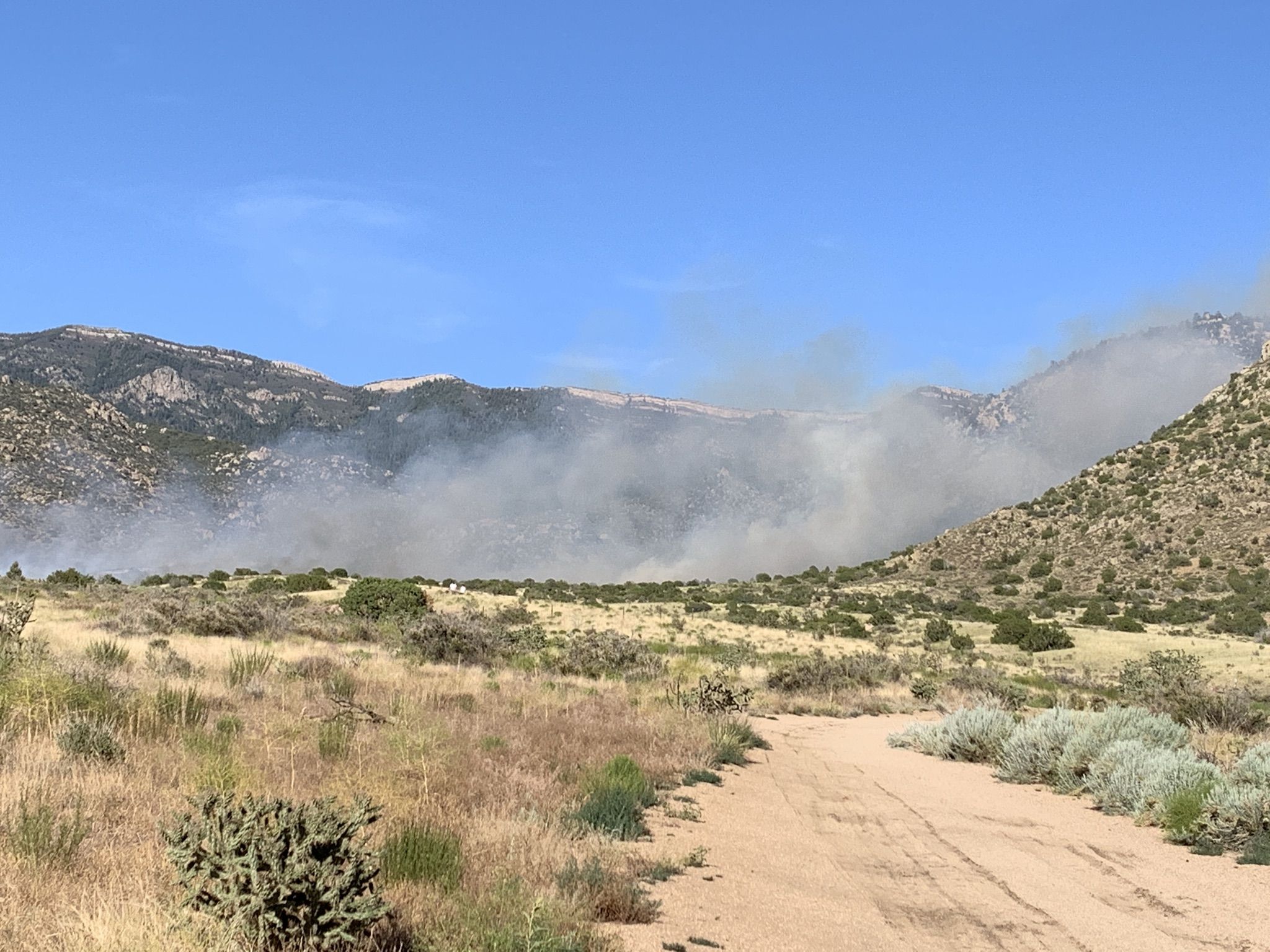 Smoke rising from distant hills, dirt path in foreground, clear blue sky. - Juan Tabo Cabin Ruins – Balloon Fiesta Park loop from Los Ranchos/Journal Center