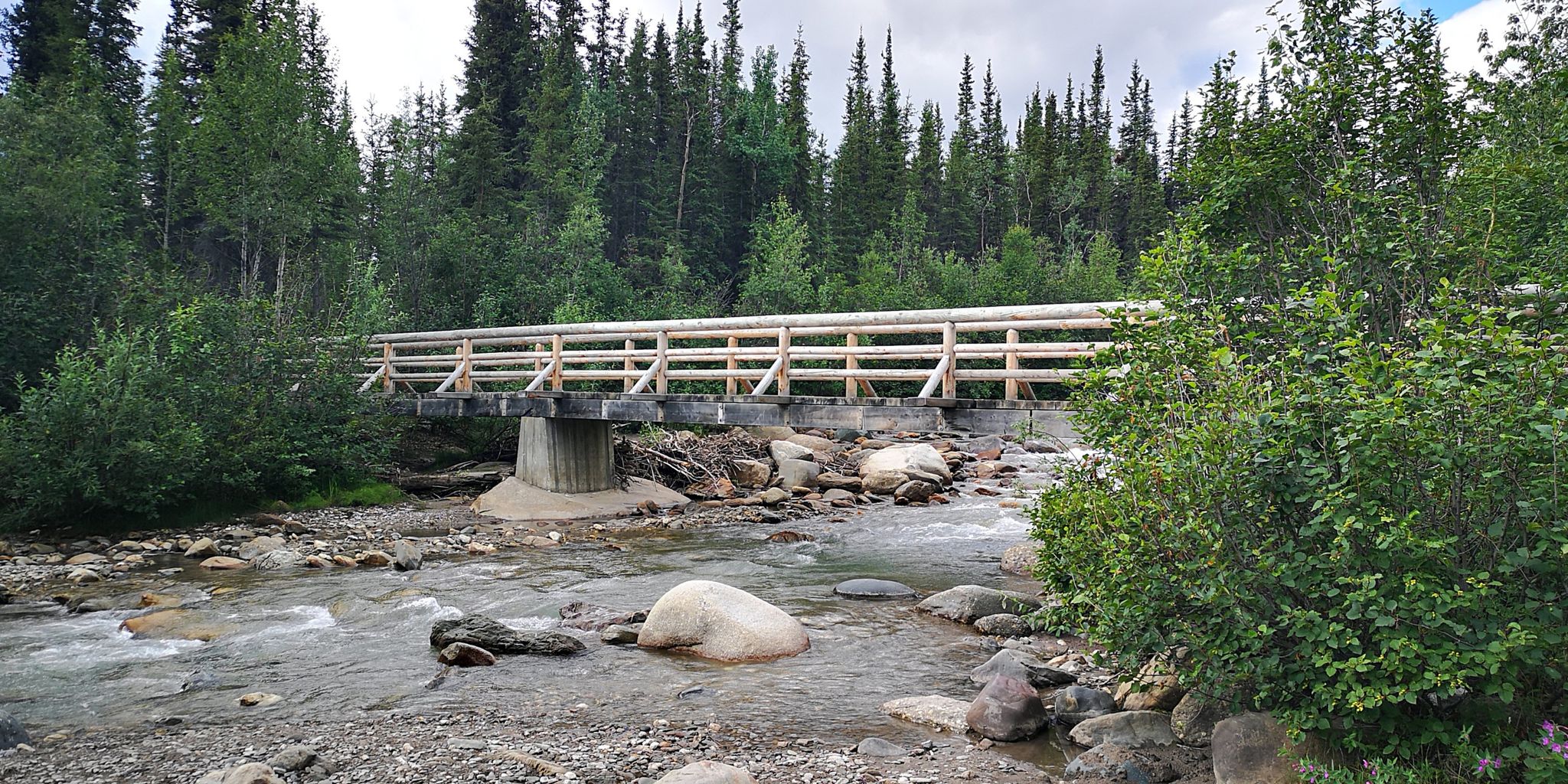 A wooden bridge with railings crosses a rocky river surrounded by green trees. - McKinley Station and Morino Loop Trail