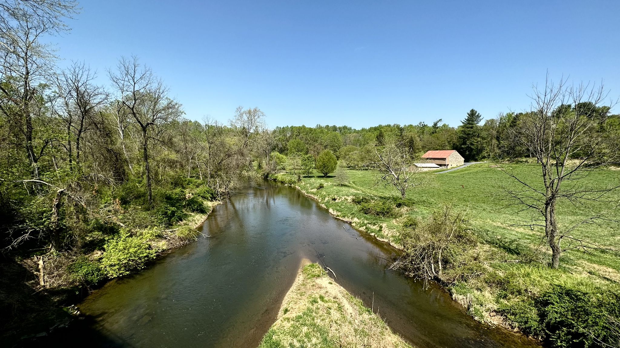 A calm river splits, flowing through green fields and trees under a bright sky. - Torrey C. Brown Rail Trail