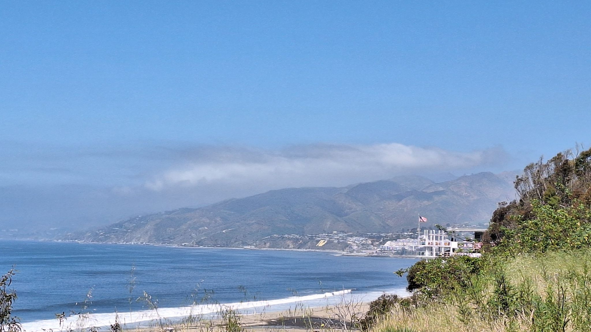 Coastal view with ocean, sandy beach, distant mountains, and a building. - Santa Monica Bluffs Trail