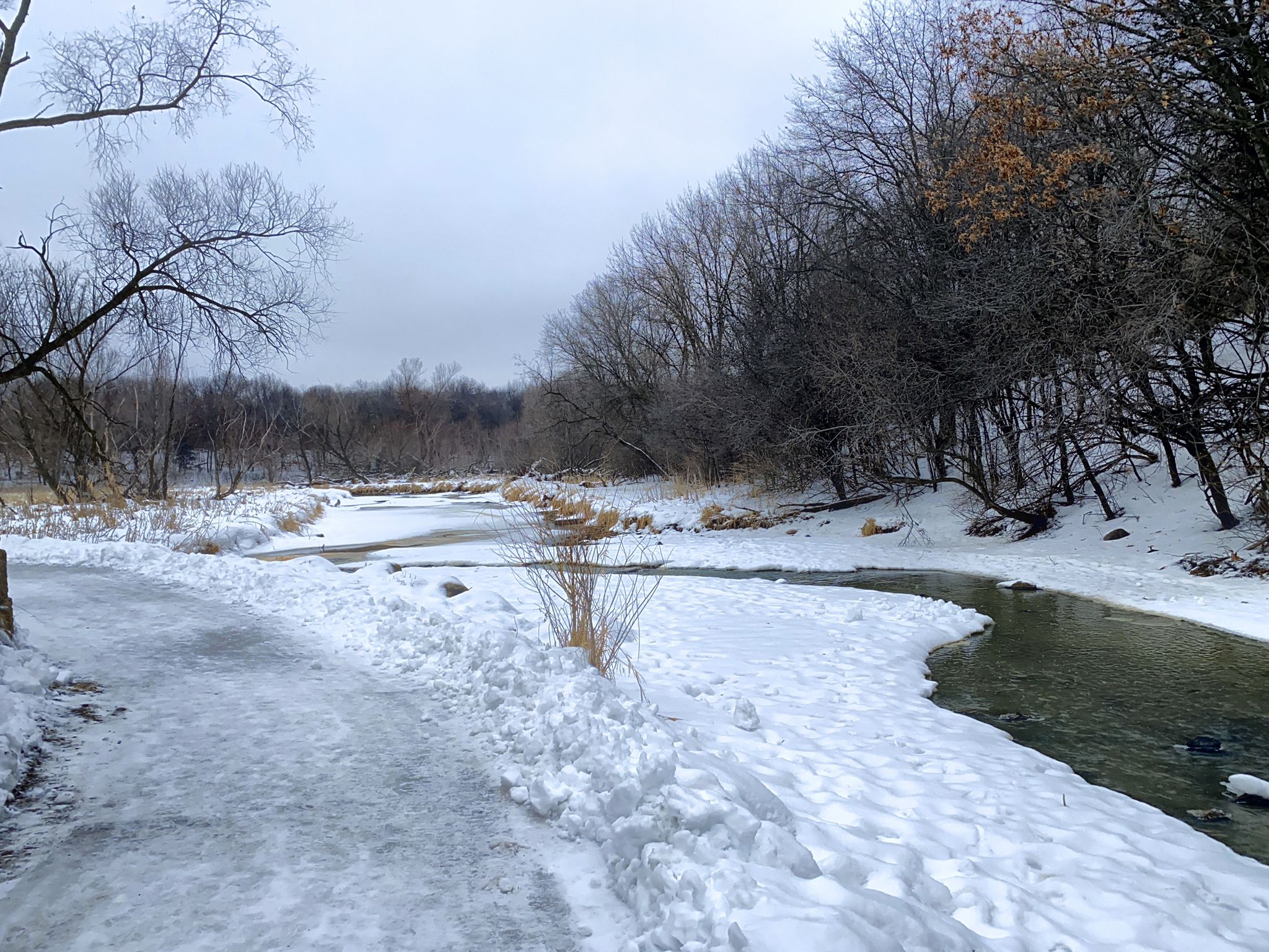 A snowy path beside a partially frozen river with bare trees under a gray sky. - Shady Oak Lake – Lone Lake Park Trails loop from Golden Valley