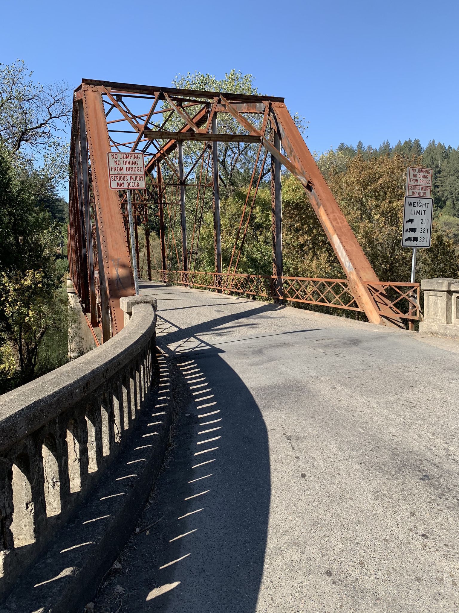 A rusty metal truss bridge with a concrete railing, bathed in bright sunlight. - Running loop from Forest Hills