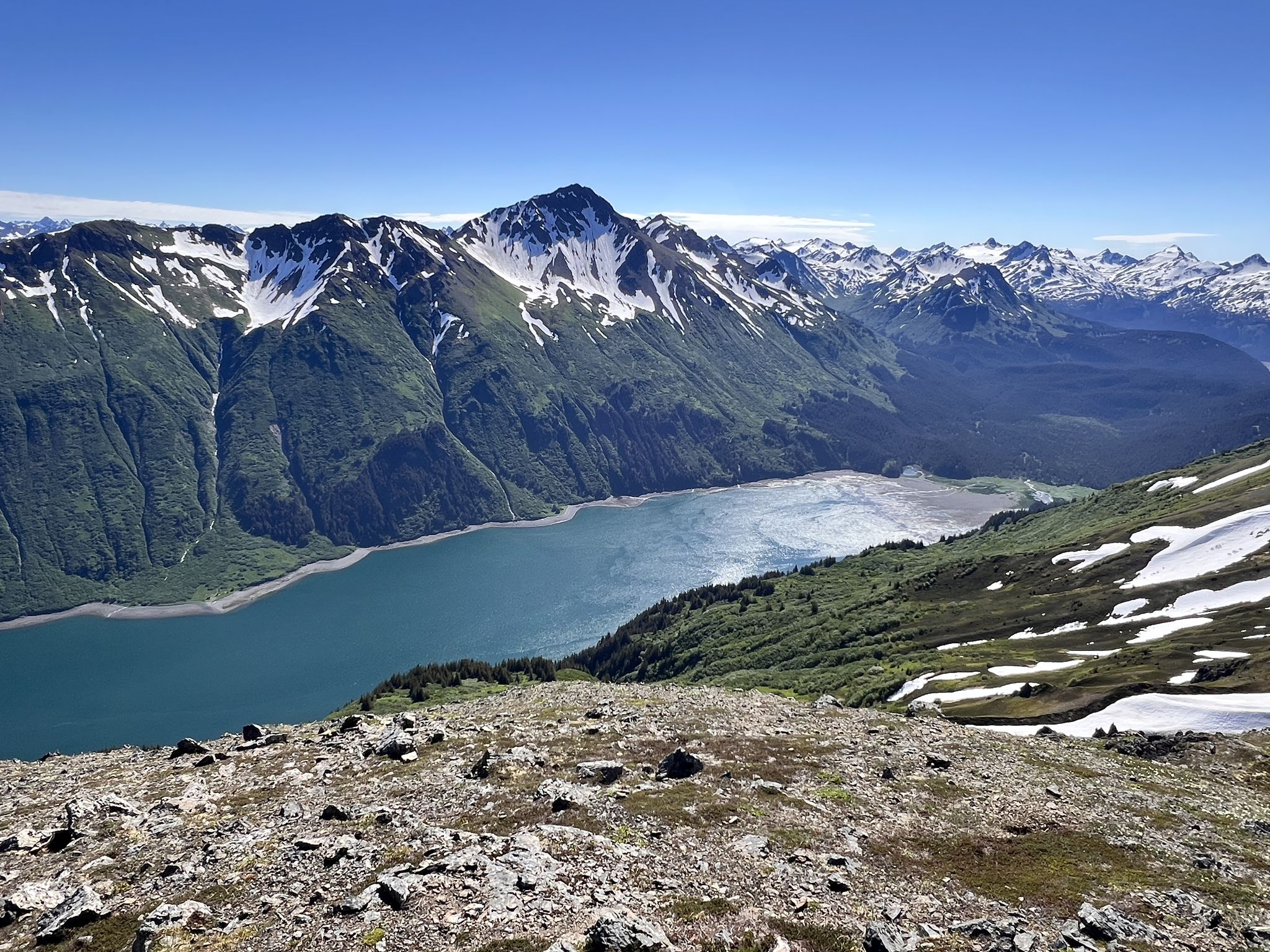 Rocky foreground, snow-capped mountains, and a blue body of water on a sunny day. - Grace Ridge Trail