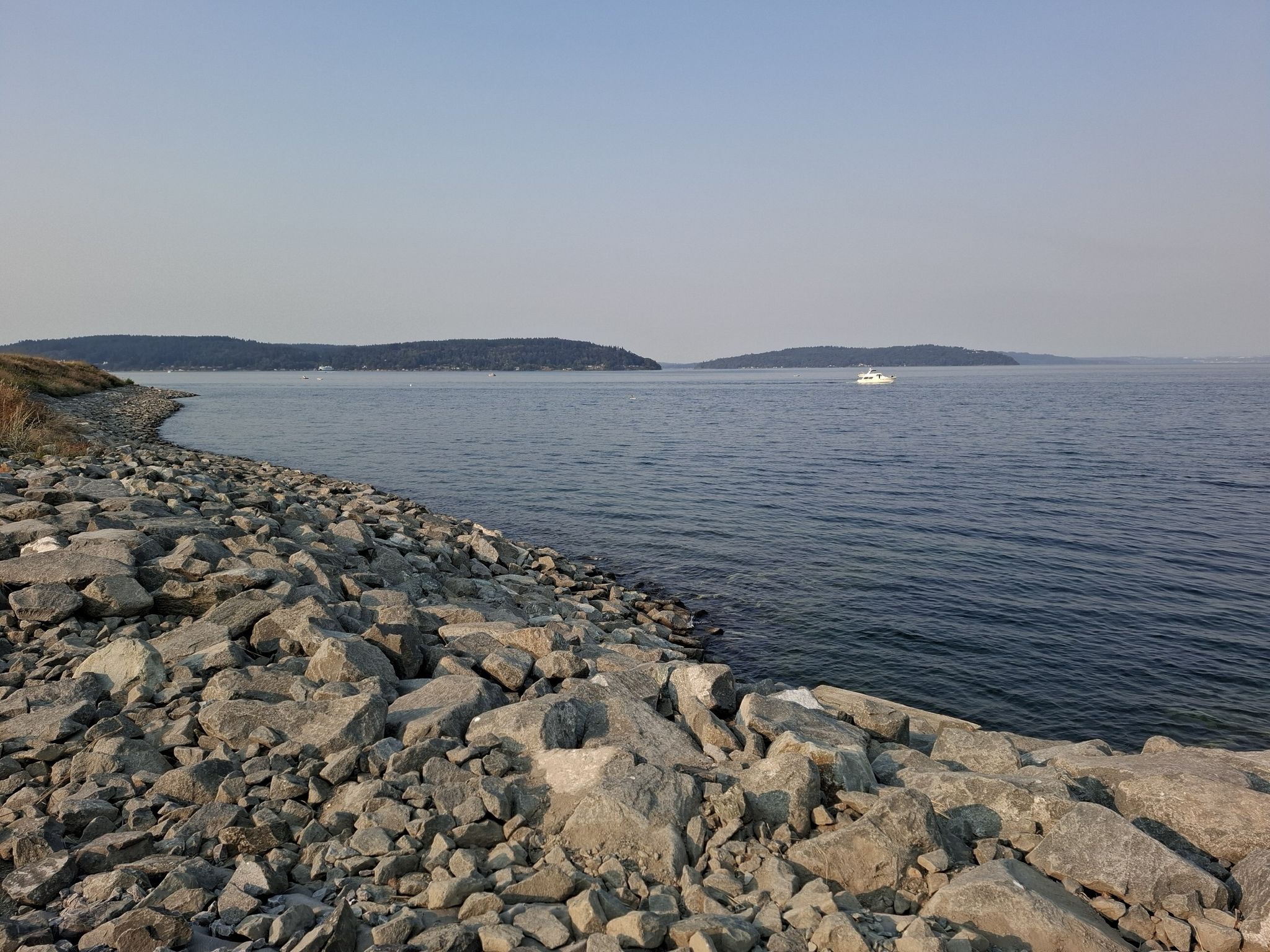 Rocky shoreline next to calm water with distant islands and a small boat. - Ruston Way Waterfront Path – Narrows Viewpoint loop from Puget Creek Natural Area