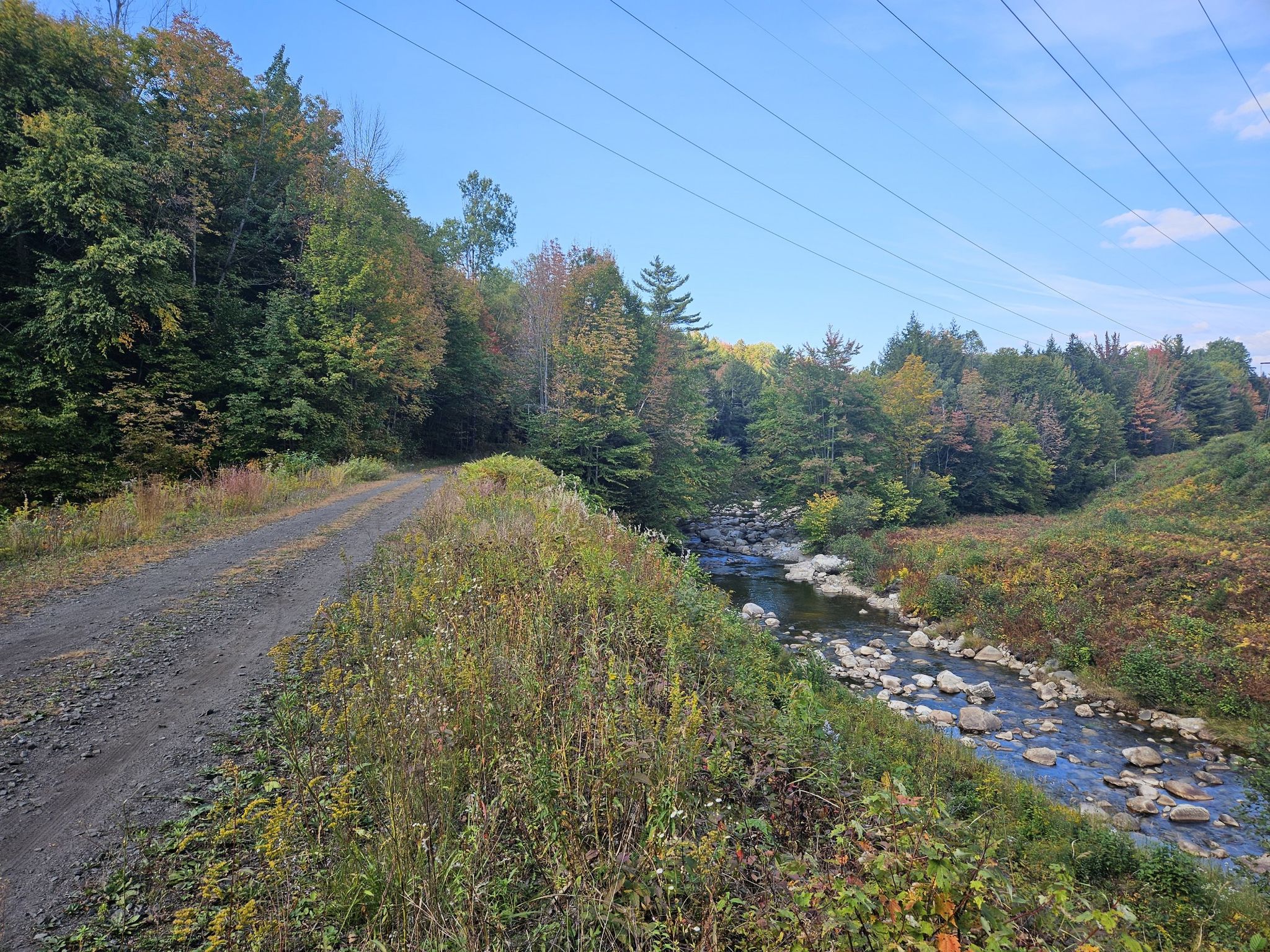 Dirt path beside a rocky stream, autumn trees, power lines. - Gravel loop from Gorham Municipal Airport