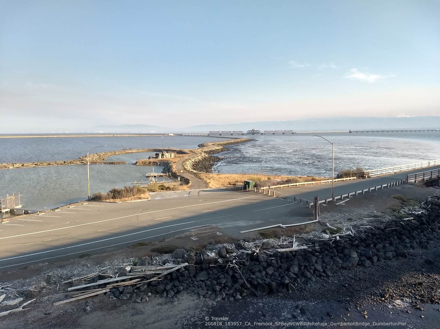 A paved road curves alongside calm water with a distant bridge and power lines. - San Francisco Bay Trail: Dumbarton Bridge