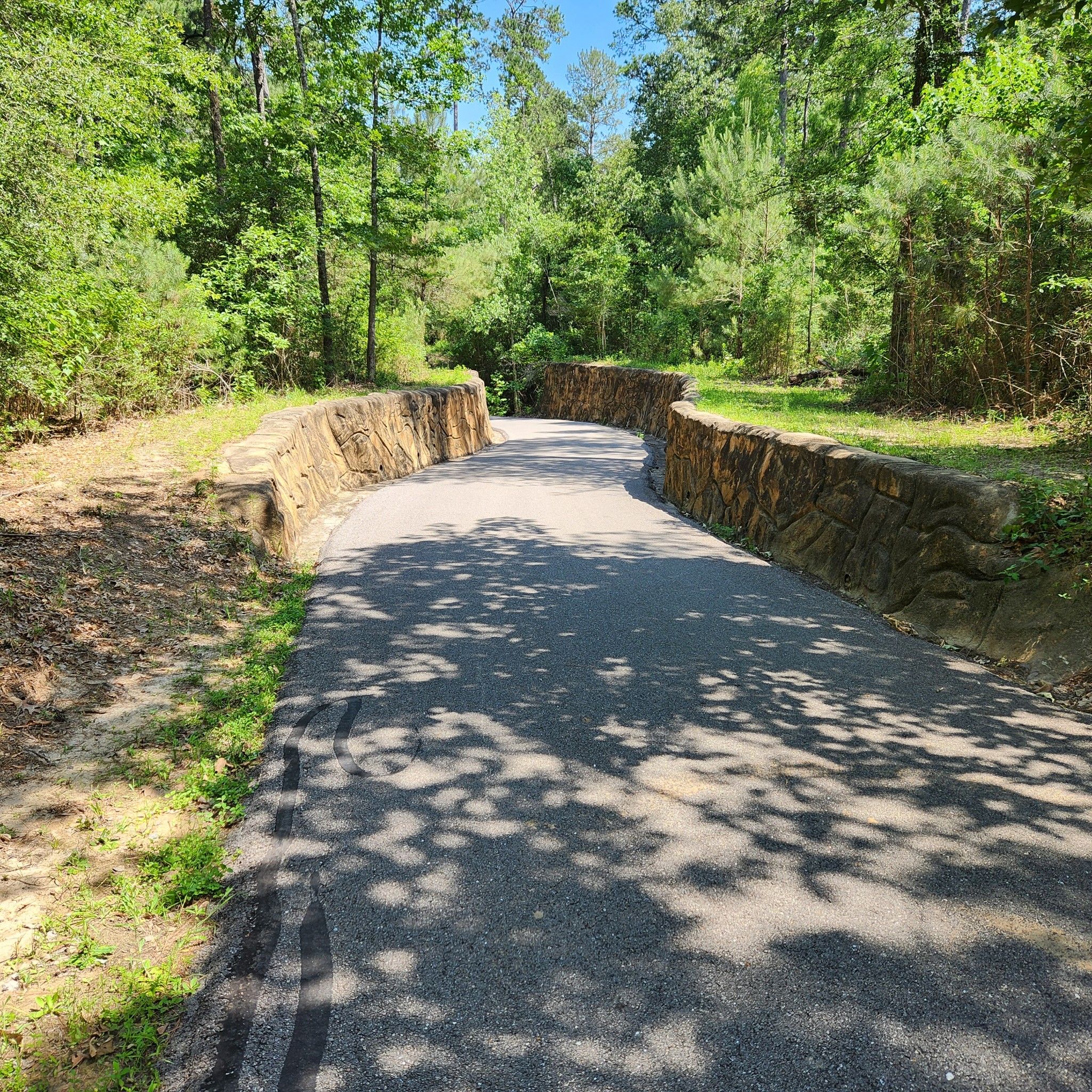 Paved path with stone walls, surrounded by green trees, under bright sunlight. - Spring Creek Greenway Trail
