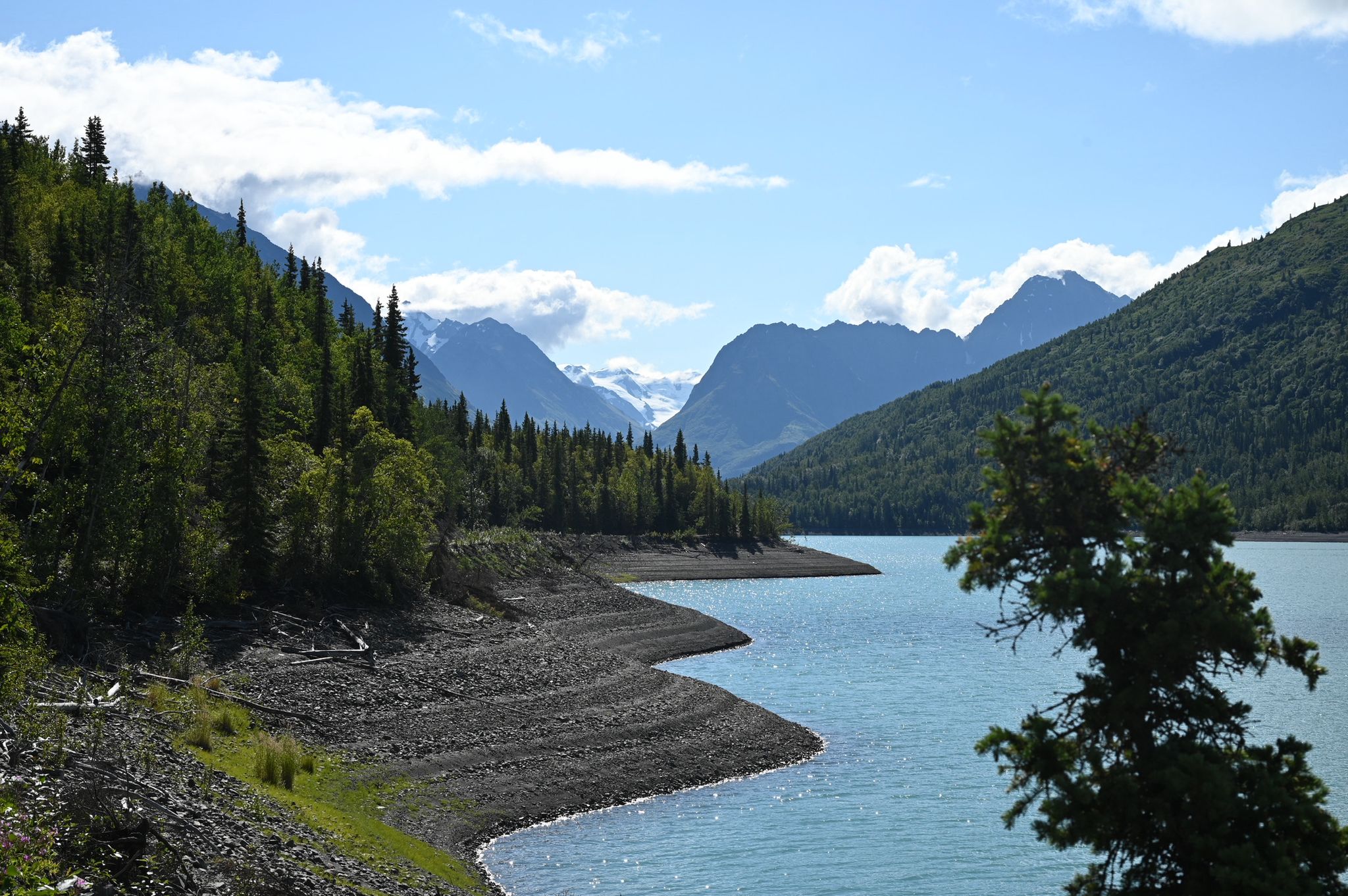 Bright daylight illuminates a blue lake, green forests, and distant snow-capped mountains. - Eklutna Lakeside Trail