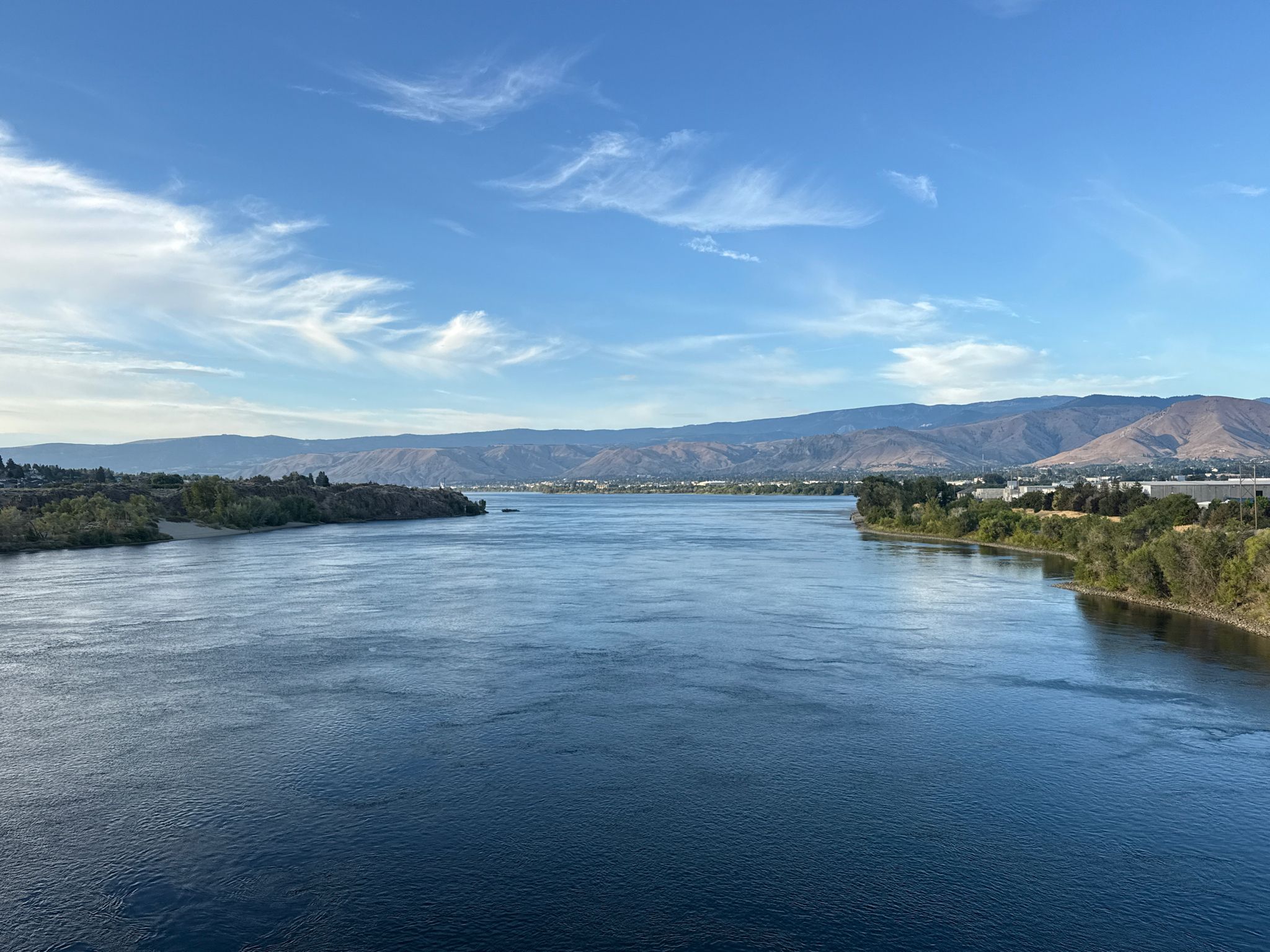 Wide river flowing between tree-lined banks, distant mountains under blue sky. - Apple Capital Recreation Loop Trail