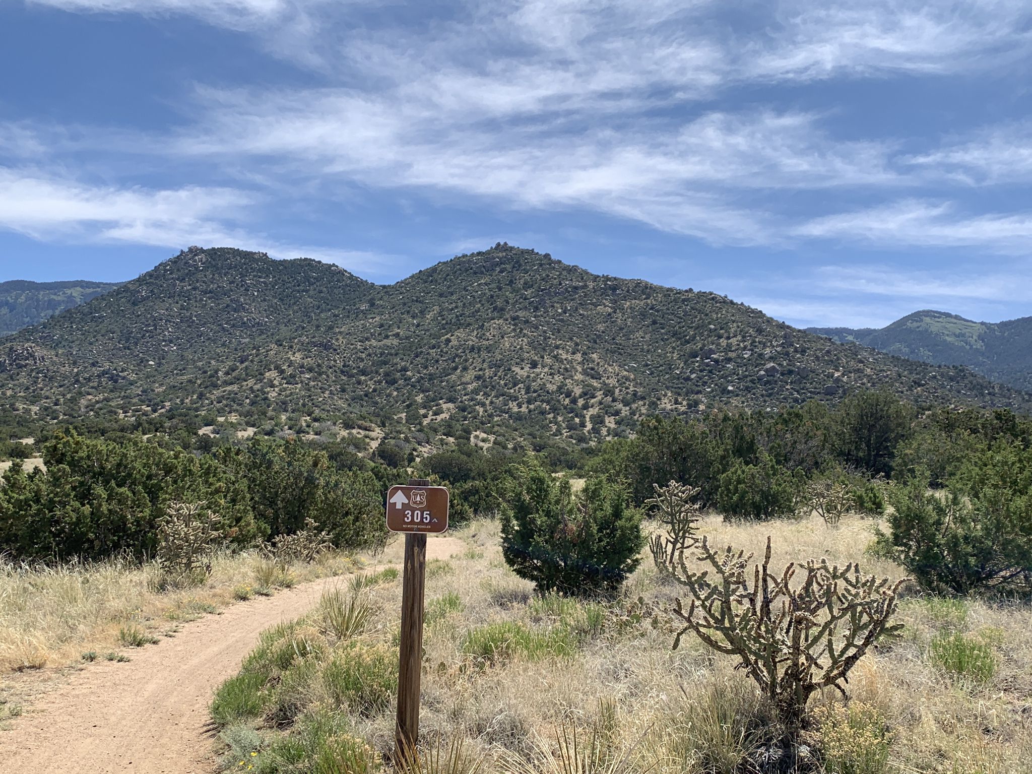 A dirt path leads to a sign, with green mountains under a cloudy sky. - Michael Emery and Trail 305 Loop