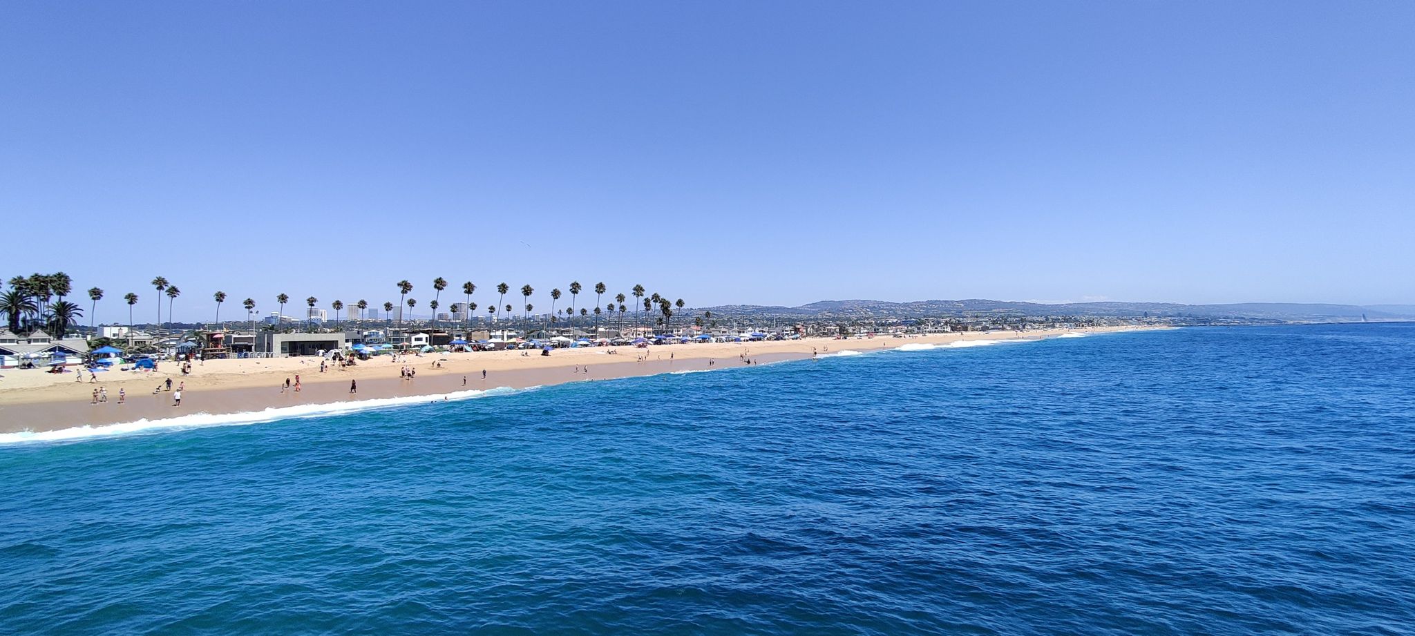 Panoramic view of a wide sandy beach with palm trees and blue ocean. - Balboa Pier & Peninsula