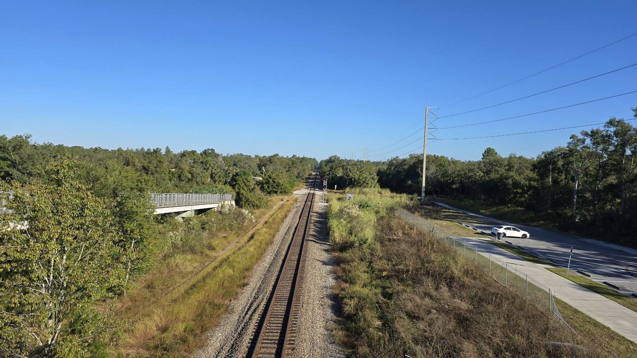 Railroad tracks, a road, and dense green trees under a clear blue sky. - Railway Overpass – Blue Spring State Park loop from DeBary