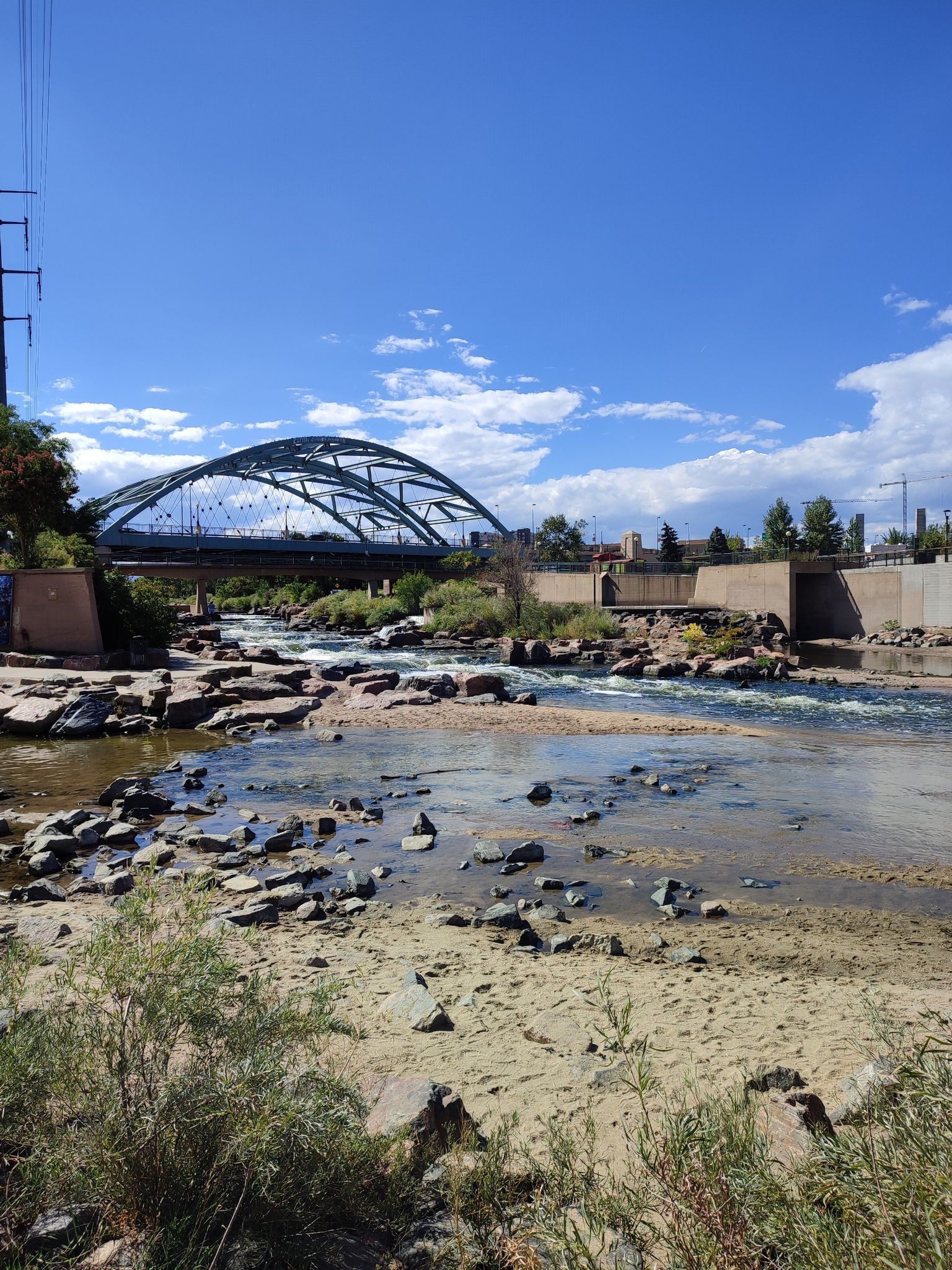 A rocky river flows under a large arched bridge on a bright, sunny day. - Cherry Creek Trail
