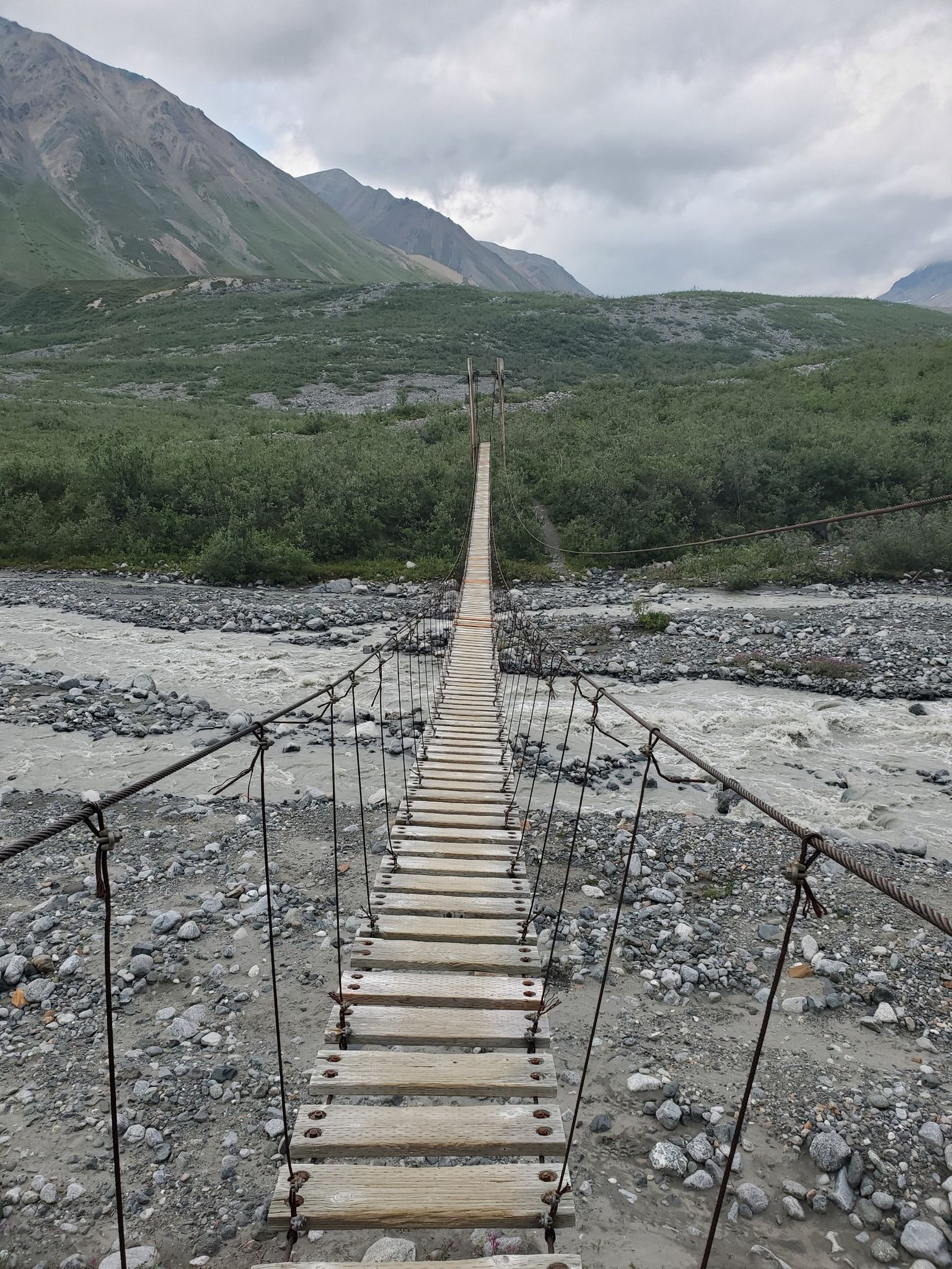 Long wooden suspension bridge over a rocky river with mountains under an overcast sky. - Gulkana Glacier Trail
