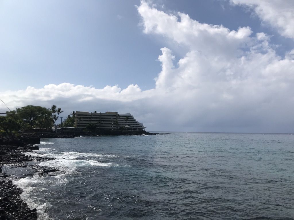 Large building on a rocky coast overlooking the ocean under cloudy skies. - Kailua Bay – Kailua-Kona loop from Kaloko-Honokōhau National Historical Park