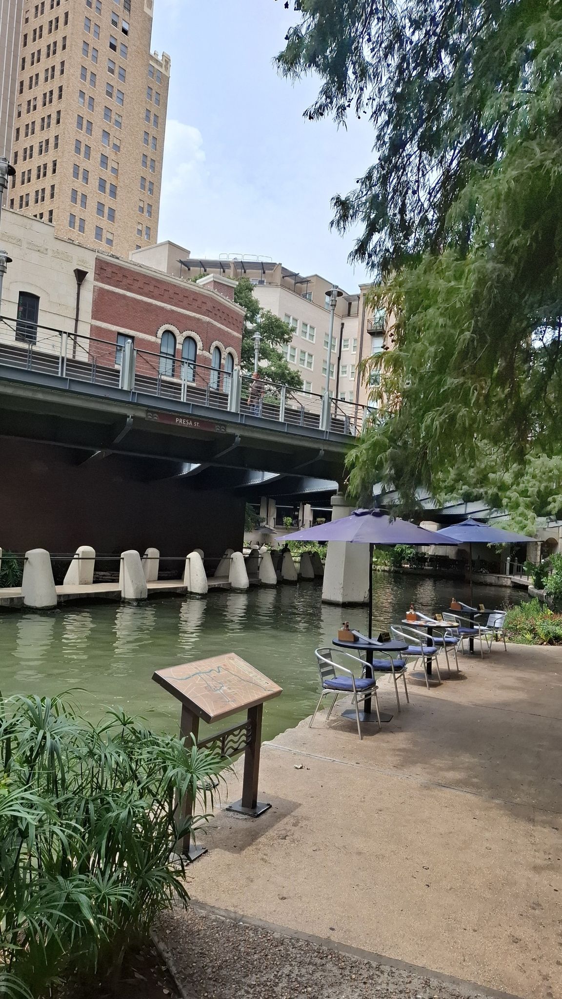 People relax at outdoor tables beside a river with city buildings and a bridge. - Eastside and Salado Creek Greenway Loop