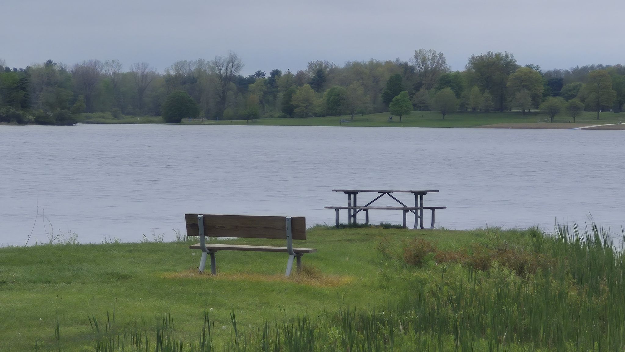 A picnic table and bench sit on a grassy bank by a wide lake. - Running loop from Banquet Tent