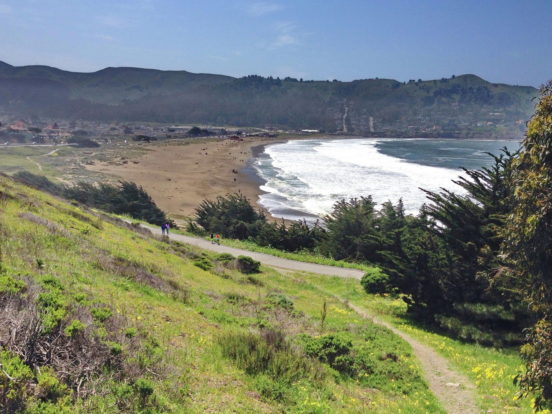 Sunny coastal view with sandy beach, green hills, ocean waves, and people walking. - Pacific Coast Bike Route: Daly City to Montara