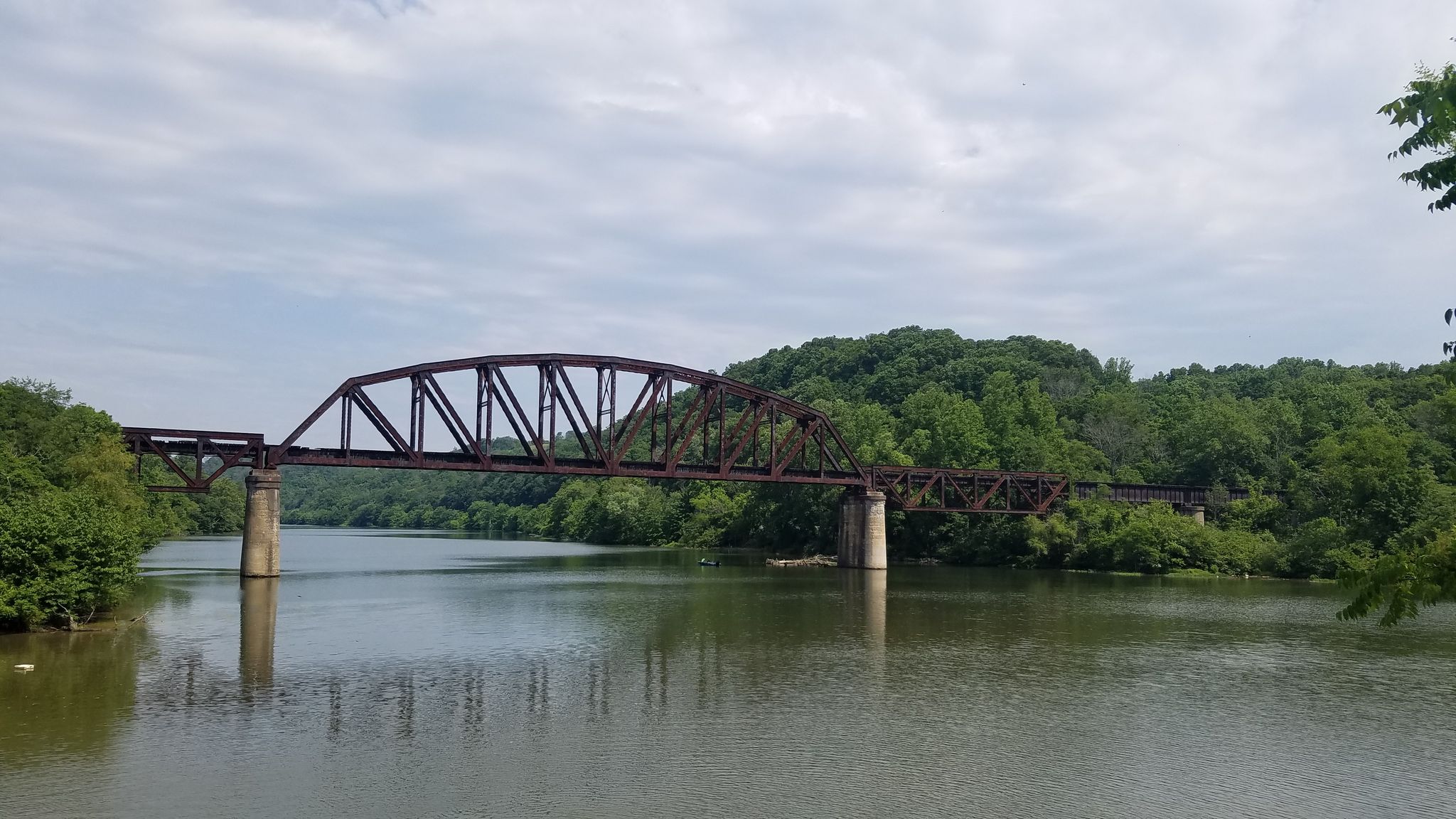 A long metal truss bridge spans a wide river surrounded by green hills. - Mon River Trail South