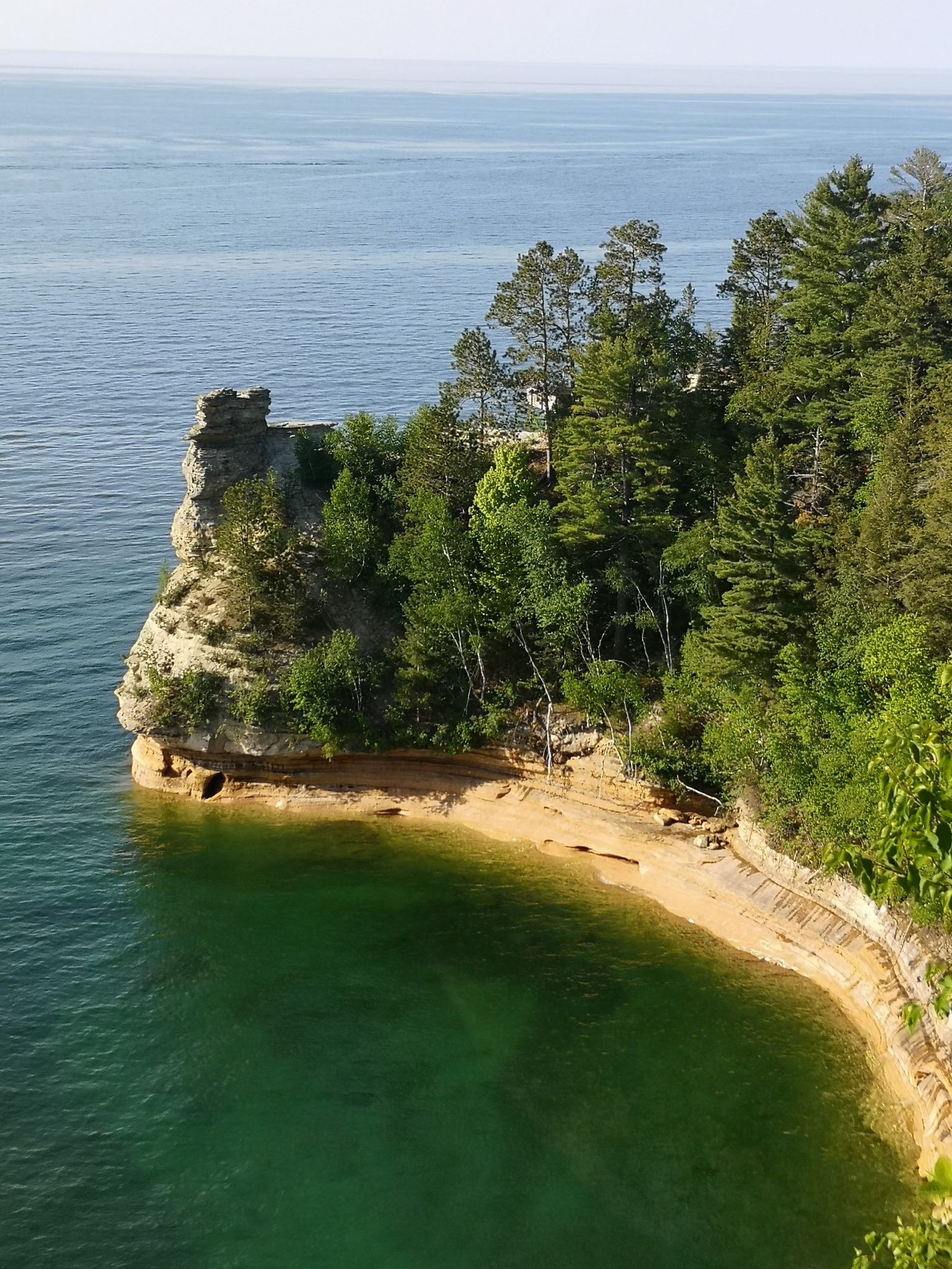 Forested cliff with a rock formation overlooking clear turquoise water on a sunny day. - Miners Castle and Miners Beach