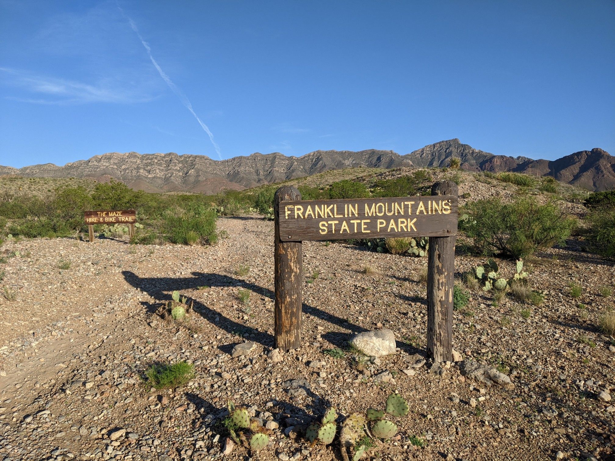 A wooden sign stands in a sunny, rocky desert with distant mountains. - Trans-Franklin Mountain Trail