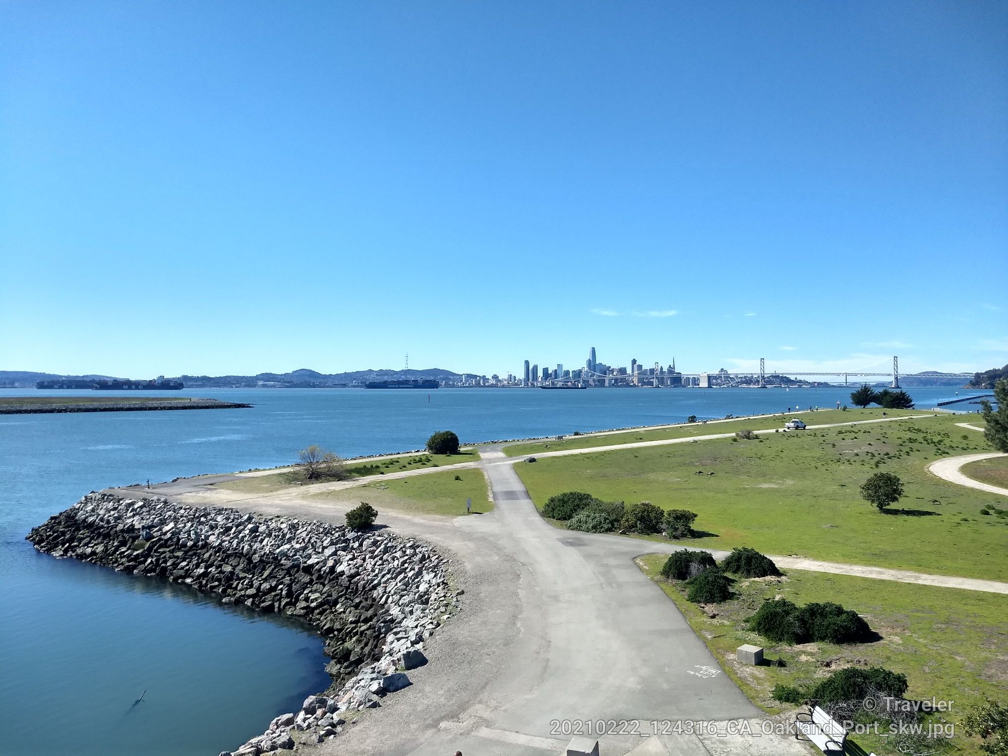 Sunny bay view with rocky shore, green land, and distant city skyline. - San Francisco Bay Trail: Middle Harbor Shoreline Park