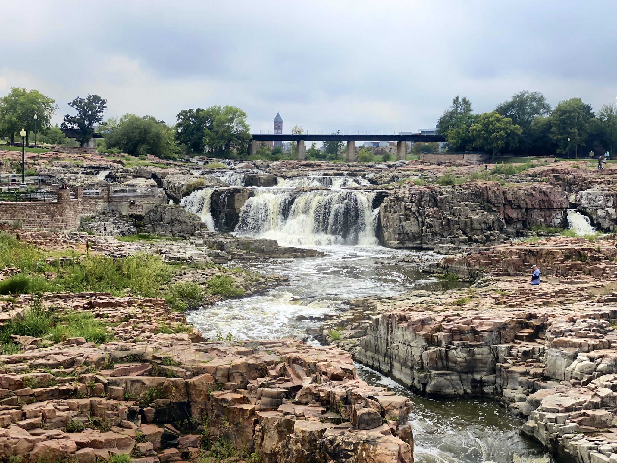 Overcast view of a wide waterfall and rocky river, with people on the banks. - The Big Sioux River Recreation Trail and Greenway