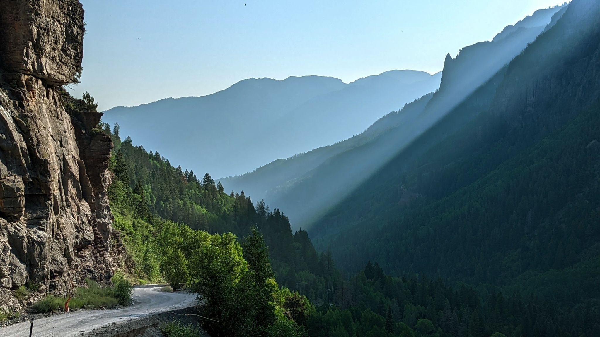 A winding dirt road along a cliff in a sunlit mountain valley. - Yankee Boy Basin OHV Route