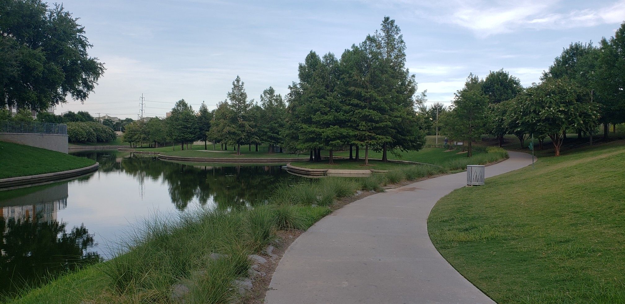 Paved path next to a calm pond, surrounded by green trees under an overcast sky. - Vitruvian Park and Brookhaven Running Track Loop