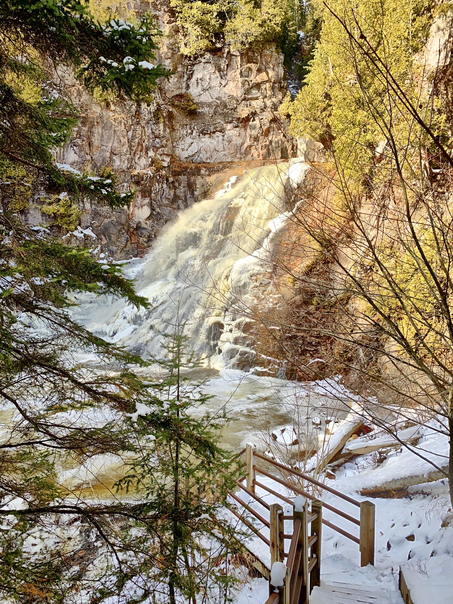 Brightly lit waterfall cascades down snowy rocks, with wooden stairs in foreground. - Caribou Falls via Superior Hiking Trail