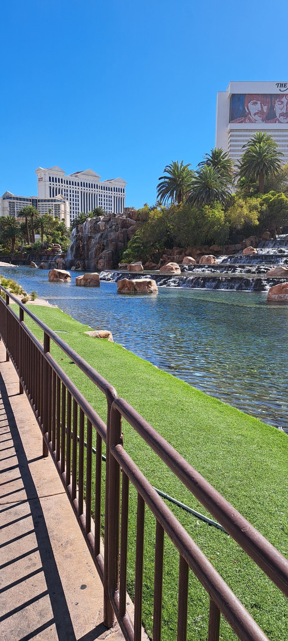 Pathway next to a canal with green turf and distant buildings. - The Strip – The Venetian Resort loop from MGM Grand