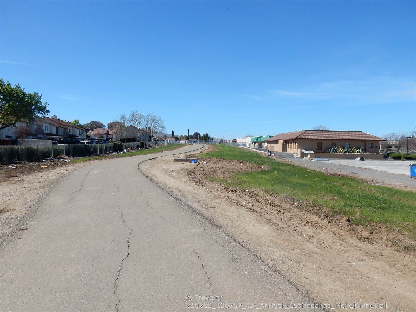 Paved path curving right, with buildings and green grass under clear sky. - Mokelumne Trail