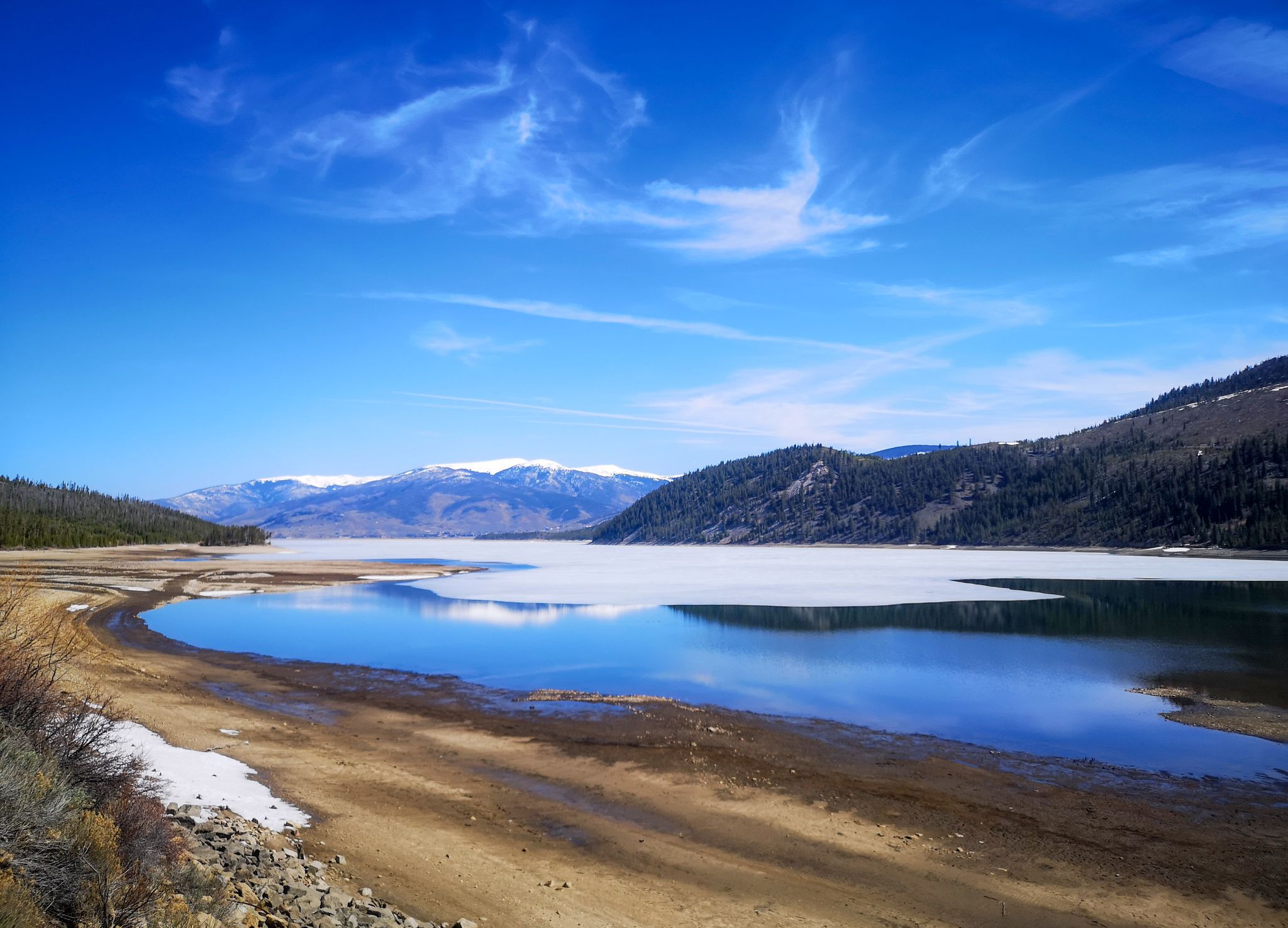 Partially frozen lake, sandy shore, snow patches, mountains under bright blue sky. - View of Dillon Reservoir – Copper Mountain loop from High School