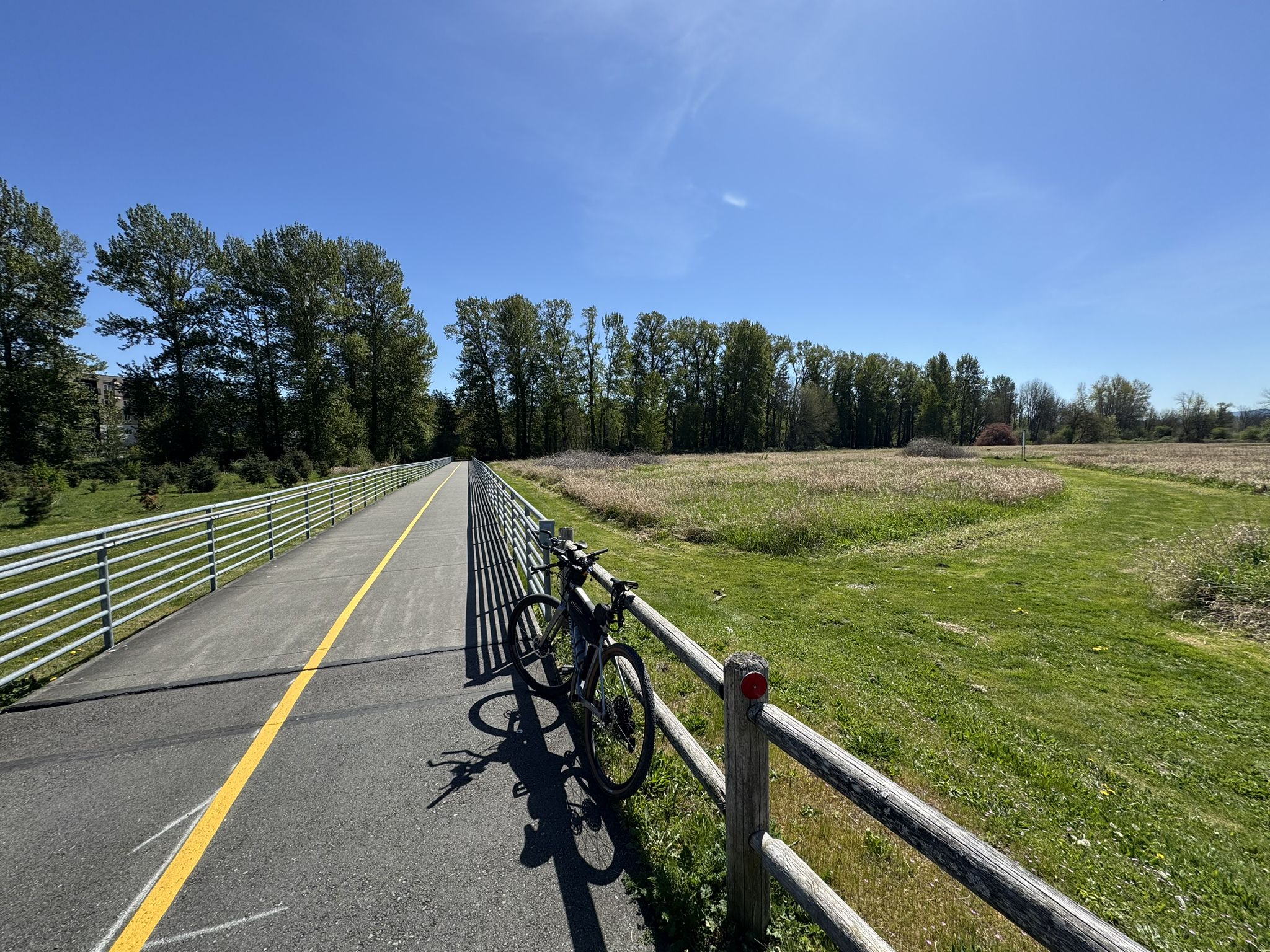 Sunny paved path with a bicycle parked by a fence, next to a grassy field. - Marymoor Connector – Sammamish River loop from Marymoor Park