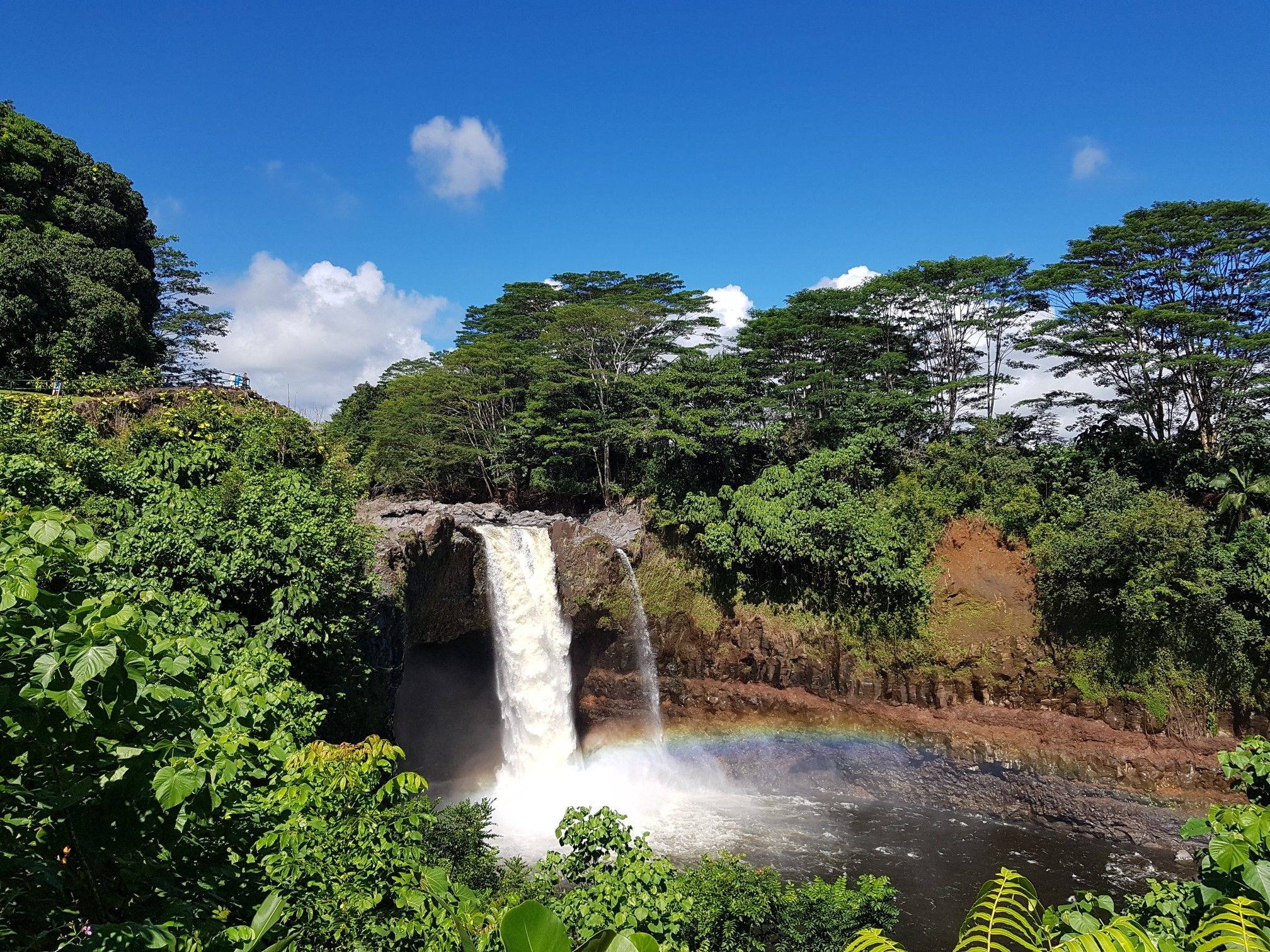 A wide waterfall cascades into a pool, with a rainbow and lush green trees. - Rainbow Falls via Kaumana Caves loop