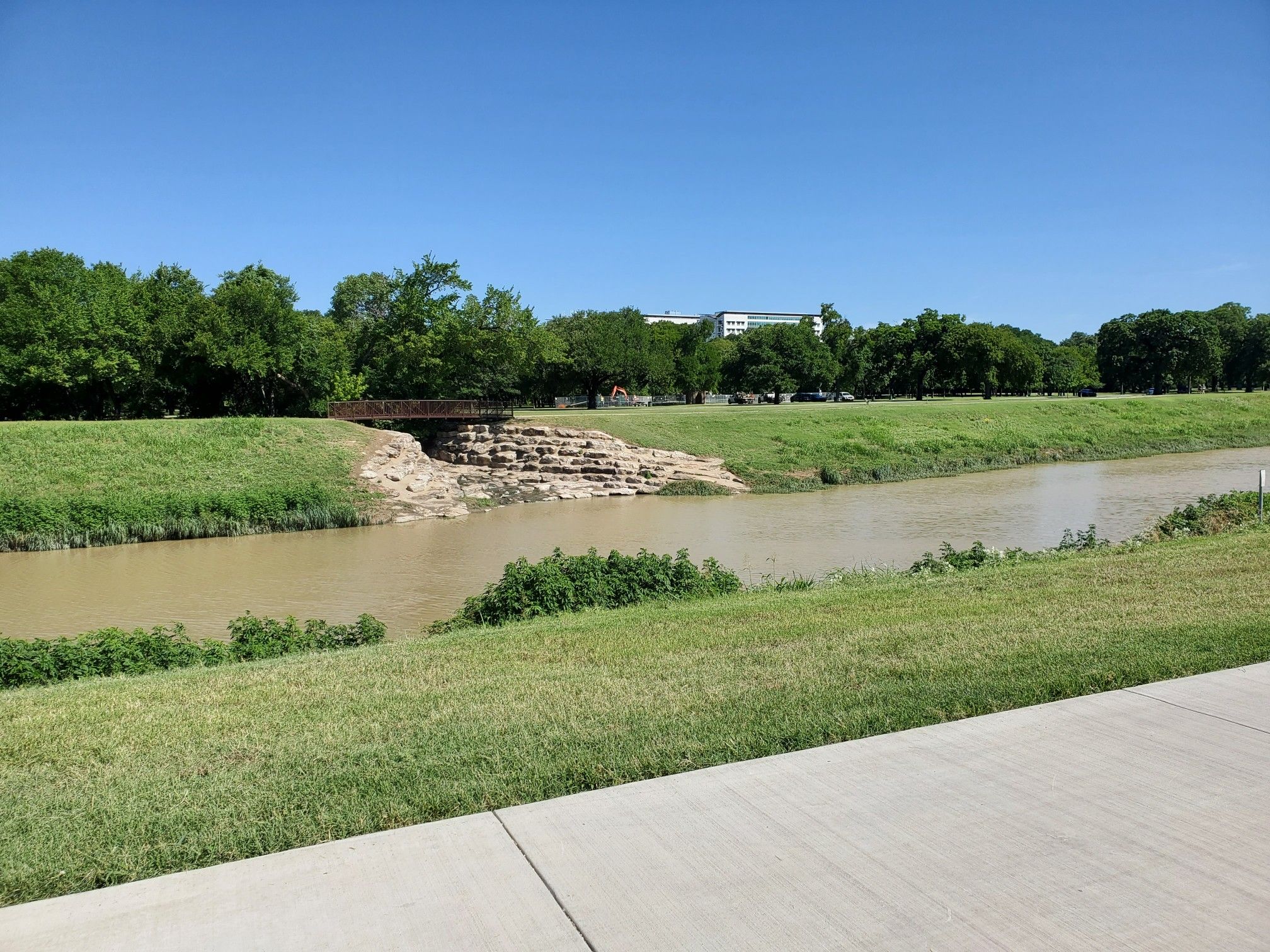 A wide river flows between green grassy banks under a clear blue sky. - Trinity Trails: Gateway Park Loop