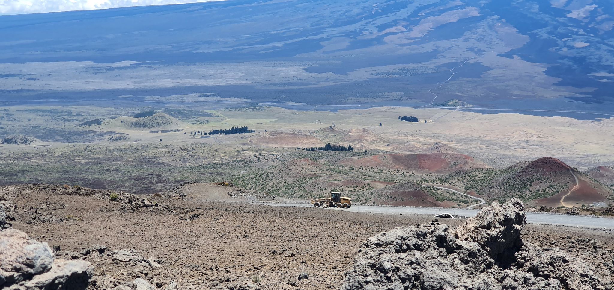 A distant vehicle drives on a road through a vast, barren volcanic landscape. - Mauna Kea summit from Puʻu Huluhulu