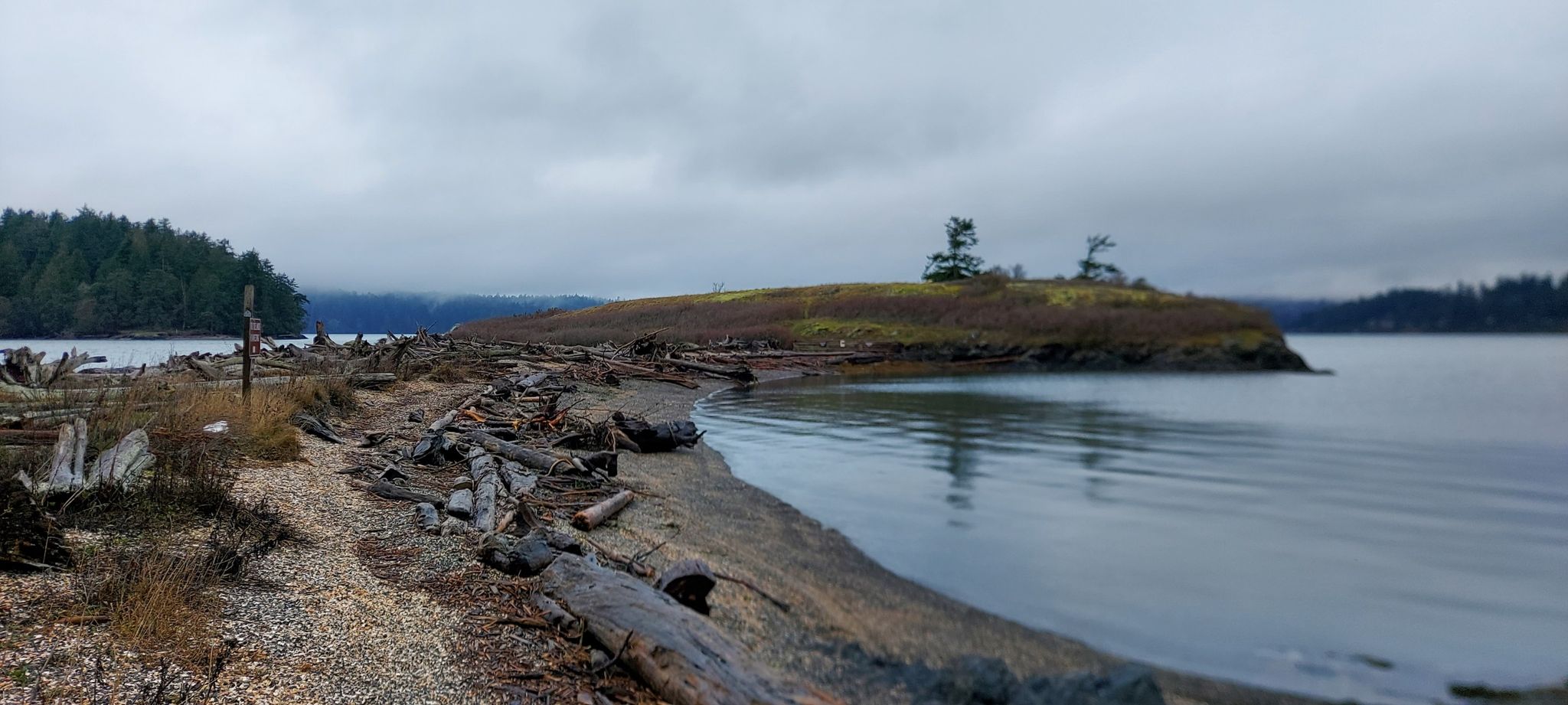 Overcast view of a narrow driftwood-covered land bridge between calm waters. - Kiket Trail loop