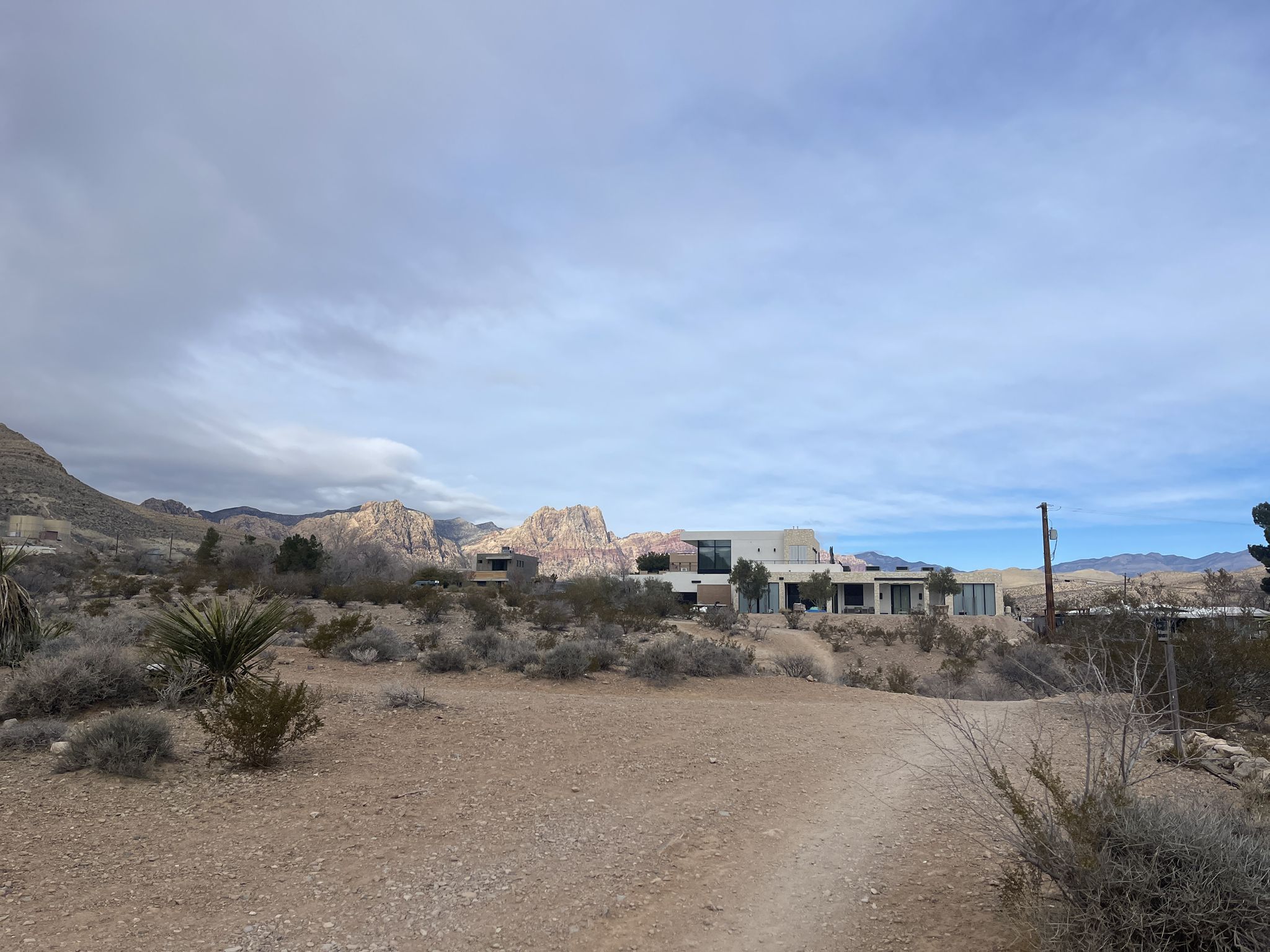 Dirt path leads towards modern buildings in a desert landscape under a cloudy sky. - Blue Diamond loop from Blue Diamond