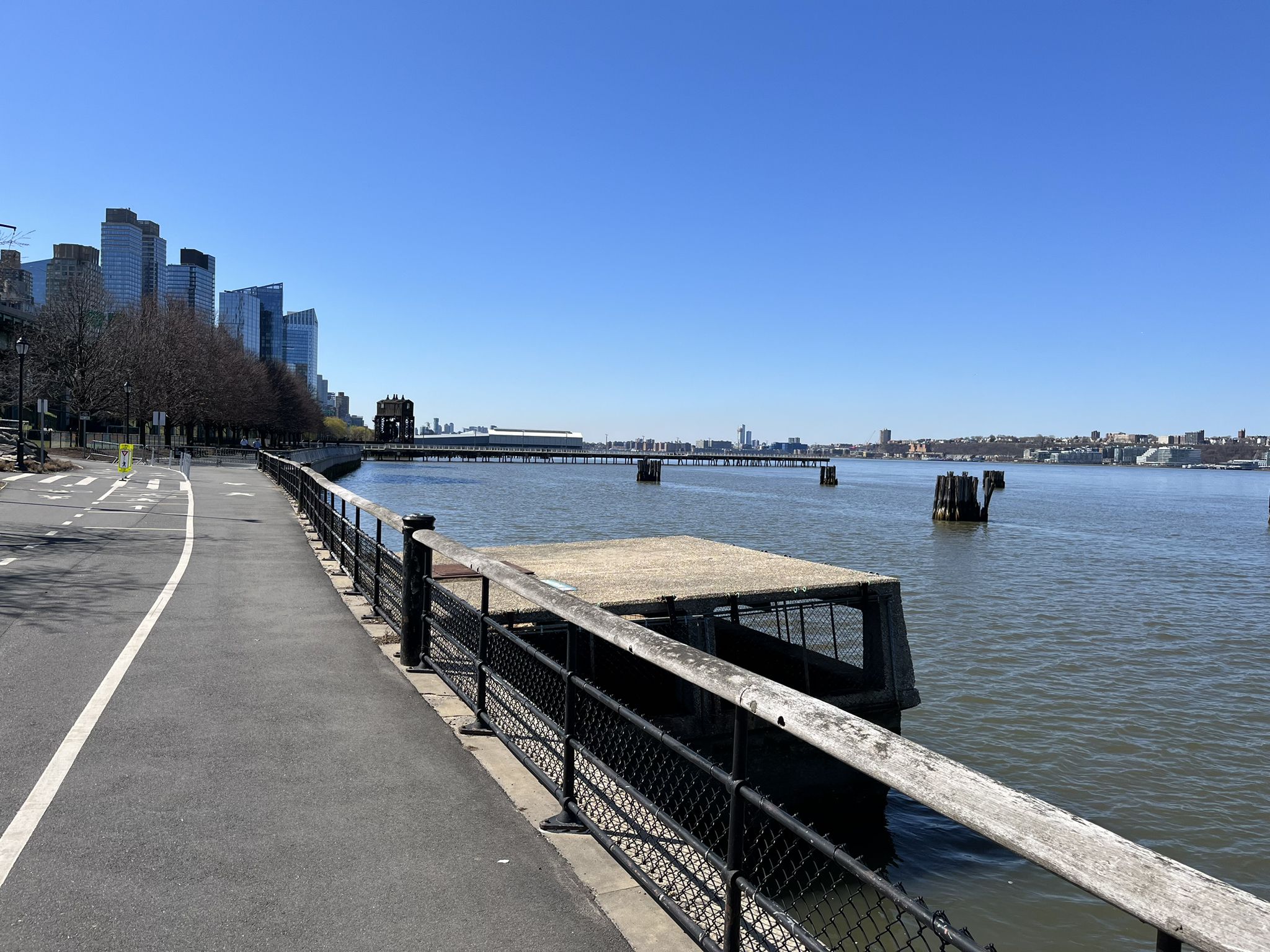 Paved waterfront path next to water with distant city skyline under clear sky. - Hudson River Greenway – Hudson River Greenway loop from 116th Street–Columbia University